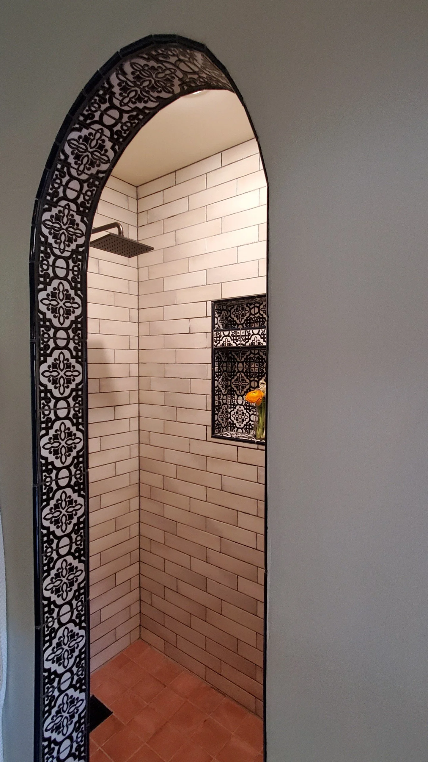 A shower space with beige subway tiles, a black decorative metal arched frame, a small black decorative shelf with a vase of orange flowers, a modern square rain showerhead, and a small floor drain.