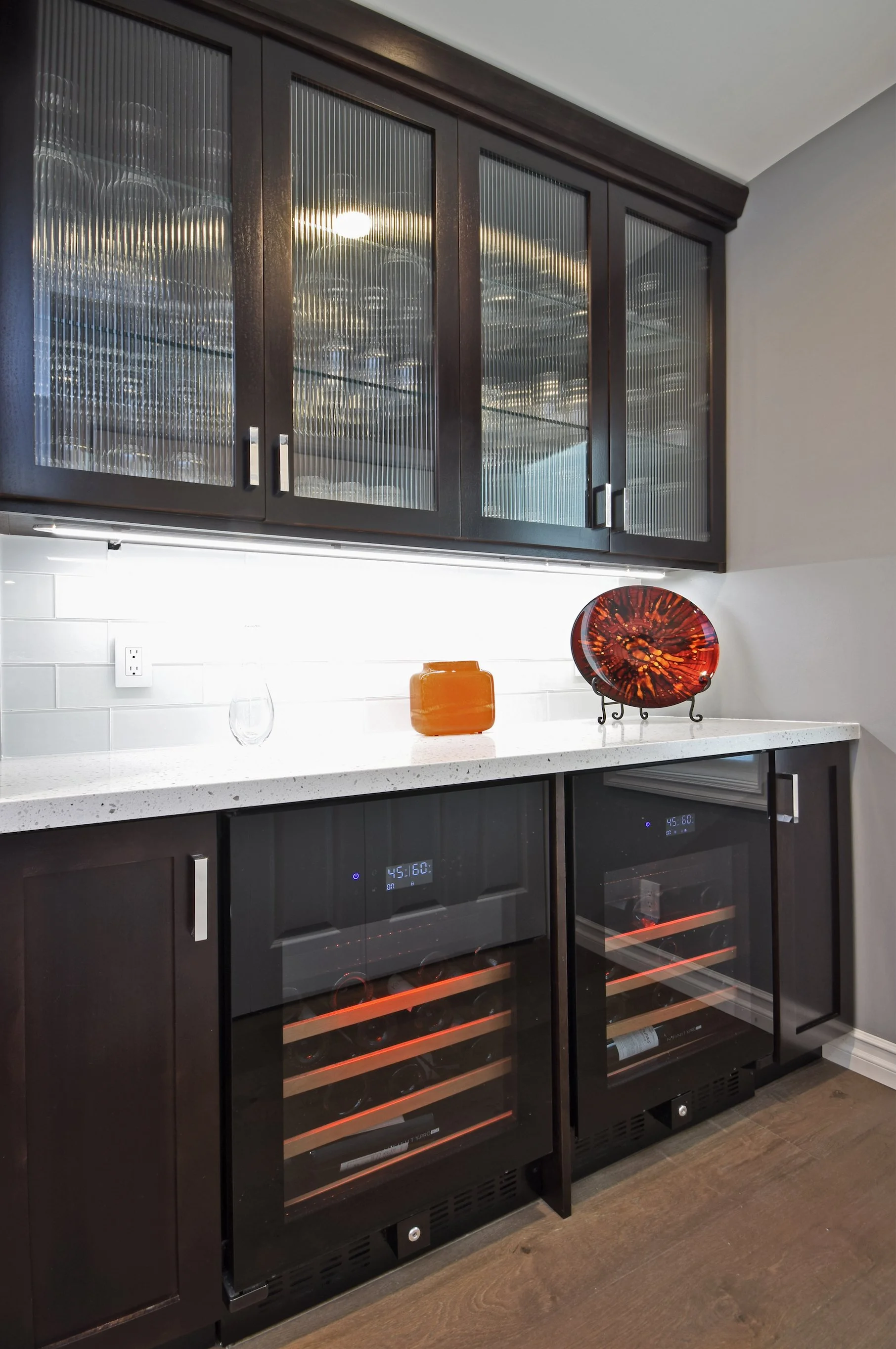 Kitchen with dark wood cabinets and glass-front wine refrigerators containing bottles of wine. A white countertop holds a decorative orange vase, a glass, and a decorative red and black plate. The wall behind has a white backsplash with an electrical