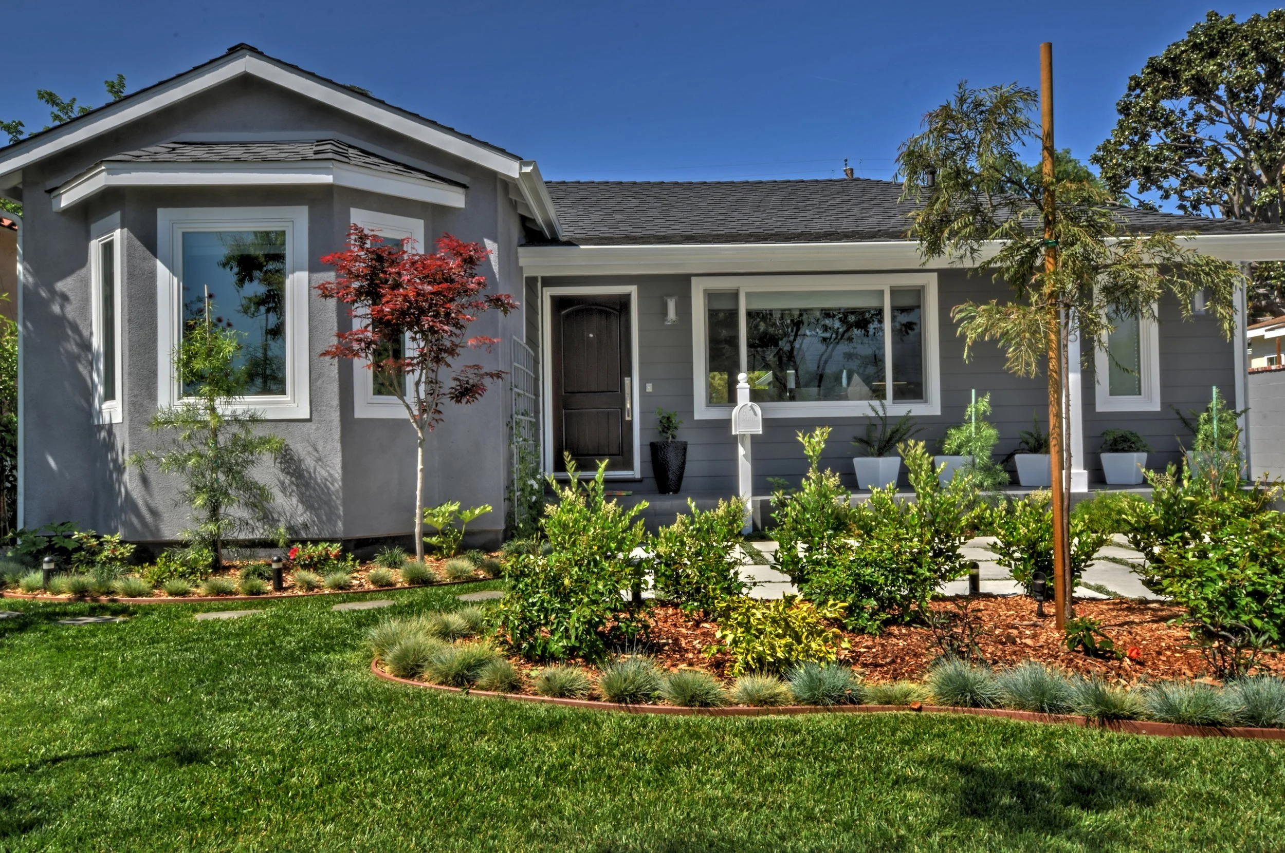 A modern house with gray exterior walls and white trim, surrounded by a lush green lawn and landscaped garden with small trees, shrubs, and potted plants, under a clear blue sky.