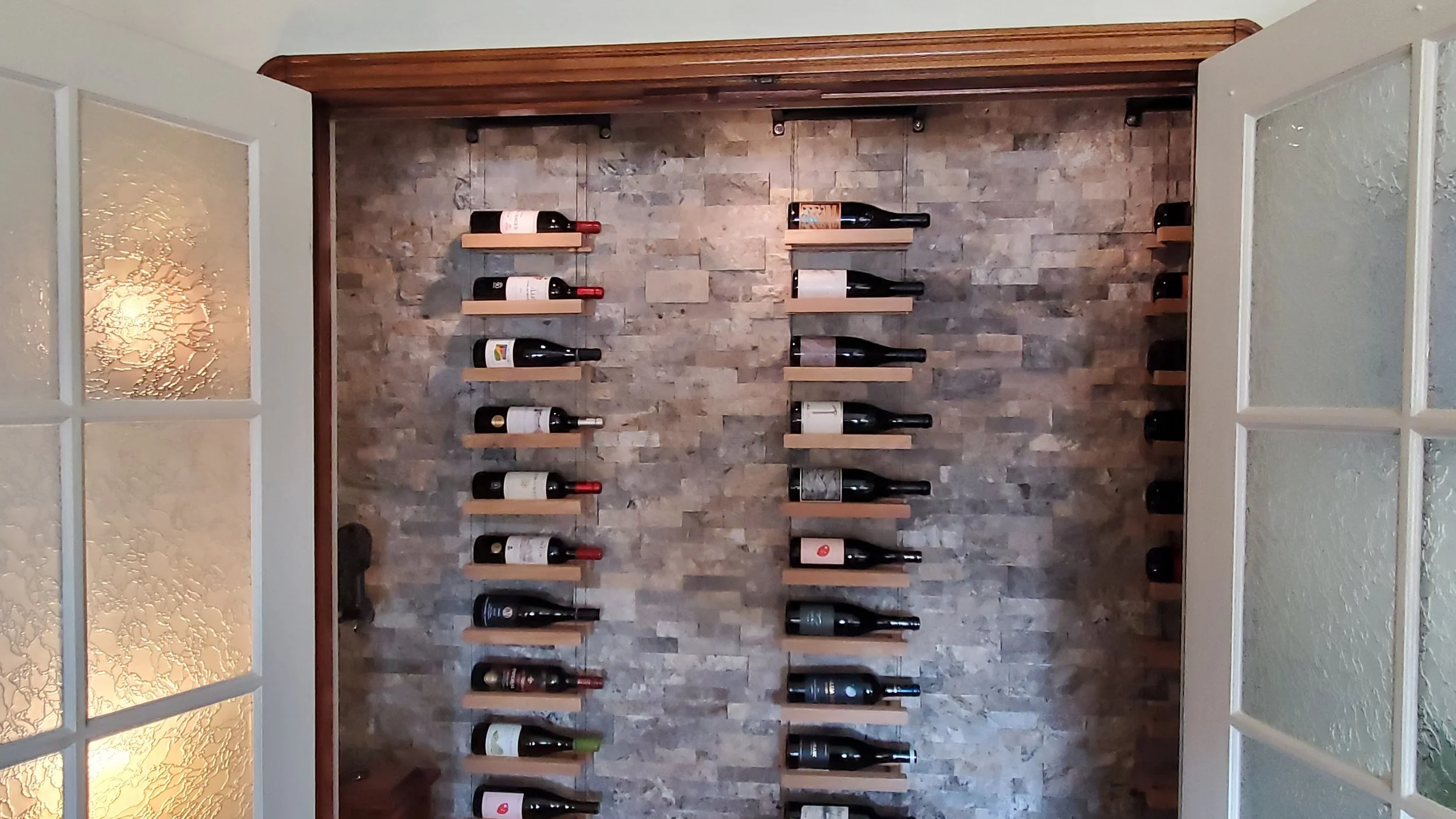 A wall with wooden shelves holding various bottles of wine, arranged in rows against a stone background, with frosted glass doors on either side.