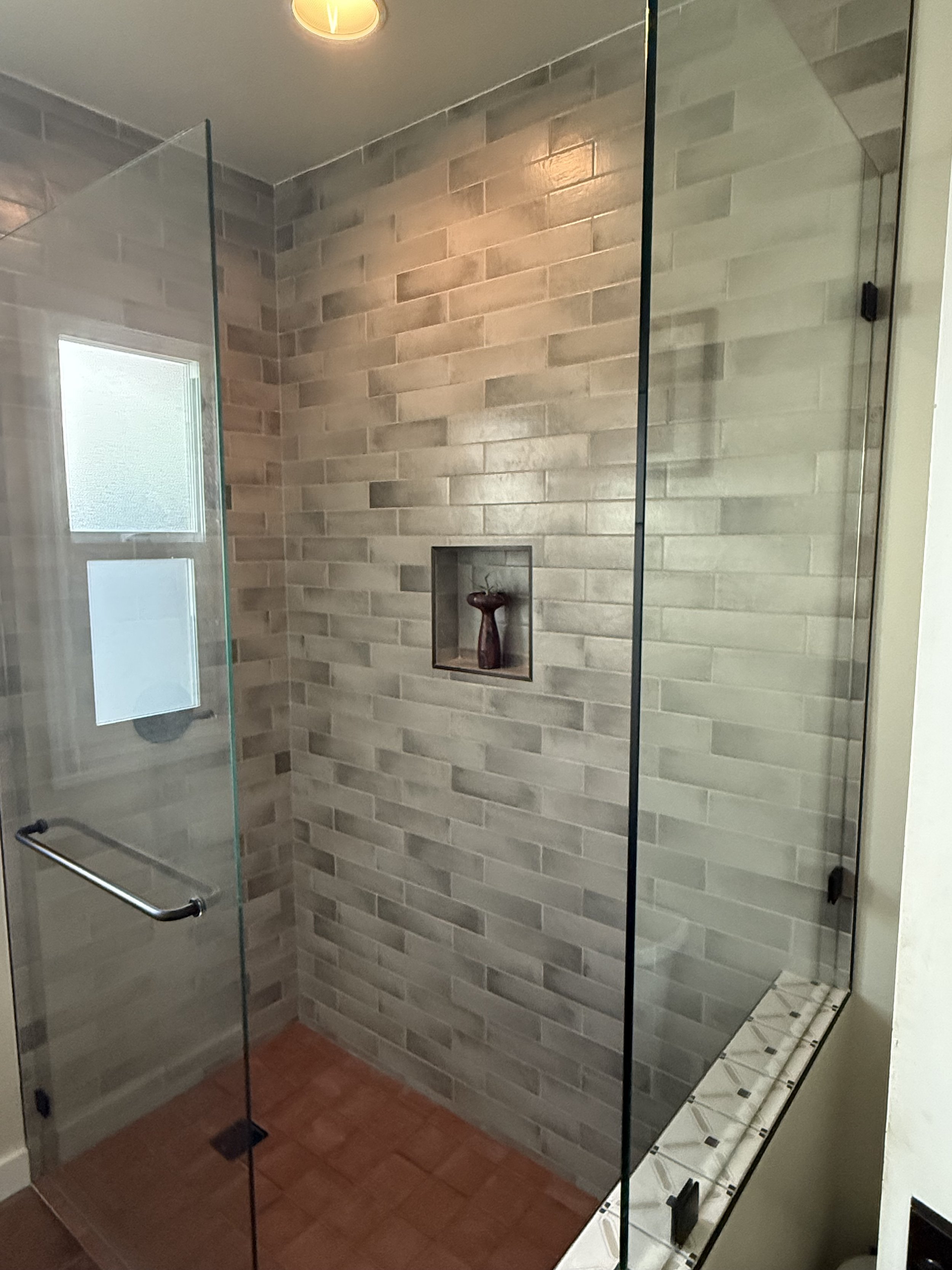 A glass shower enclosure with a brick-patterned tile shower wall, a niche with a decorative vase, a frosted window, and a floor with reddish-brown tiles.