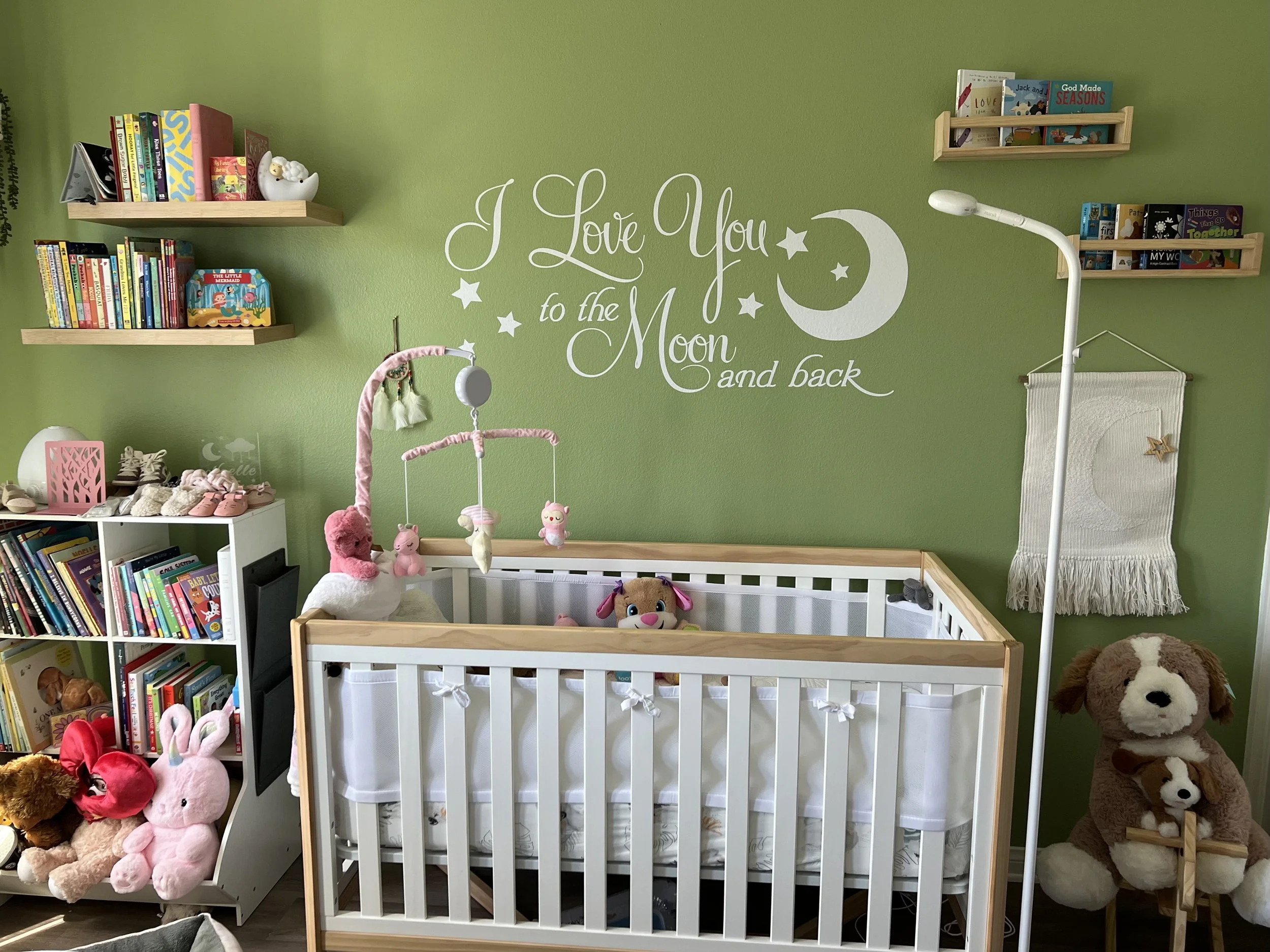 A green wall with white cursive wall decal reading 'I Love You to the Moon and back' with moon and star illustrations. Below, a white crib with stuffed animals, including pigs, a dog, and bears, is positioned beside a white book shelf filled with chi