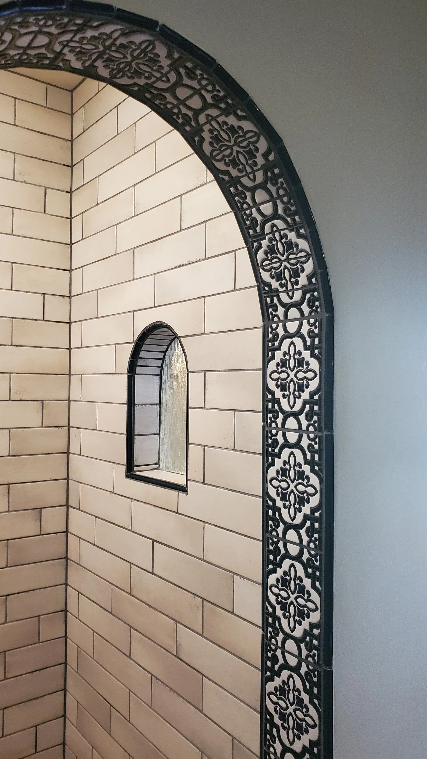 Interior of a room with beige brick wall, arched mirror, small arched window, and decorative black metal trim along the wall.