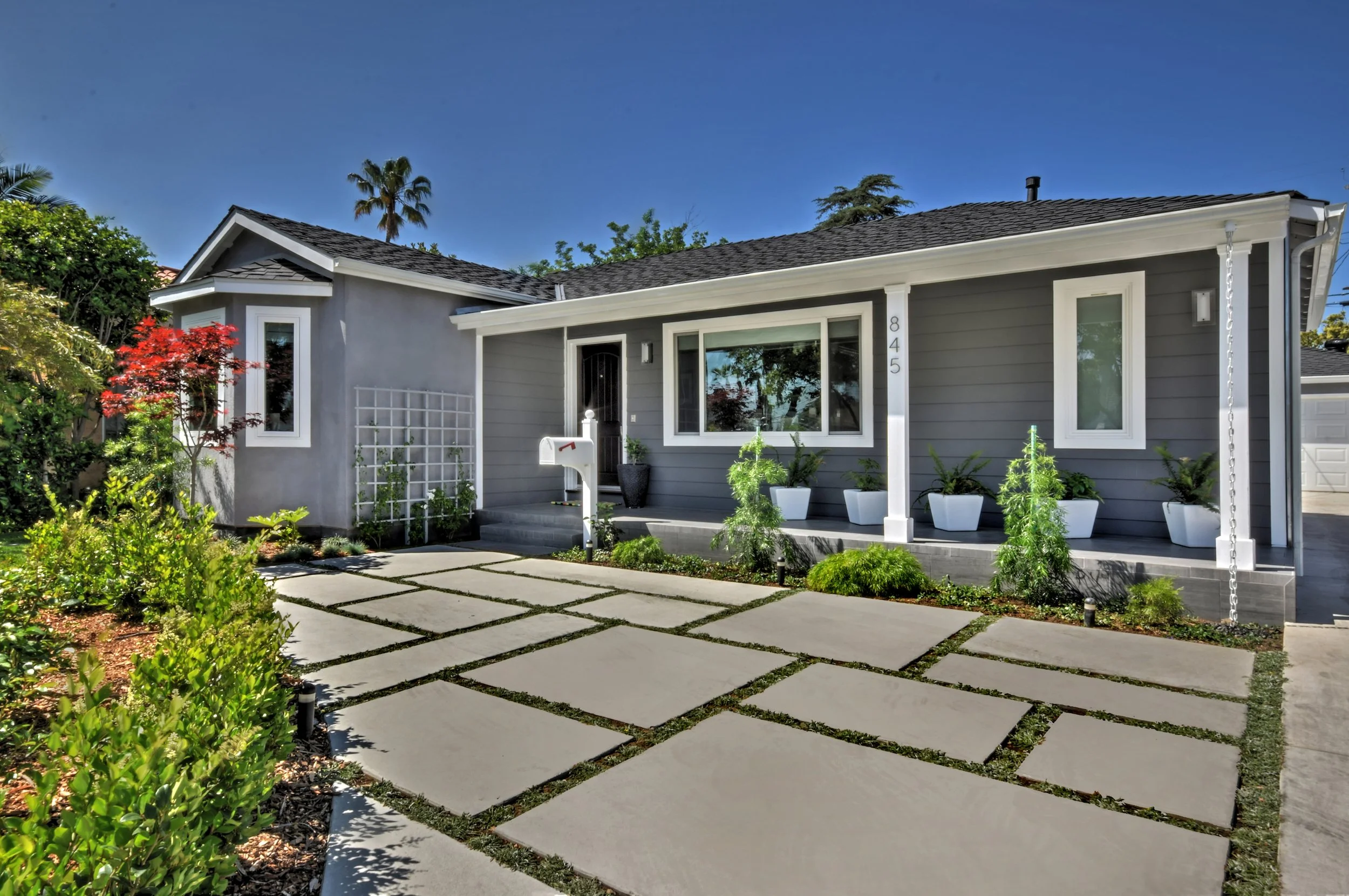 Front yard of a house with a concrete pathway, lush greenery, and potted plants, featuring a gray house with large windows, a black front door, and a well-maintained garden under a clear blue sky.