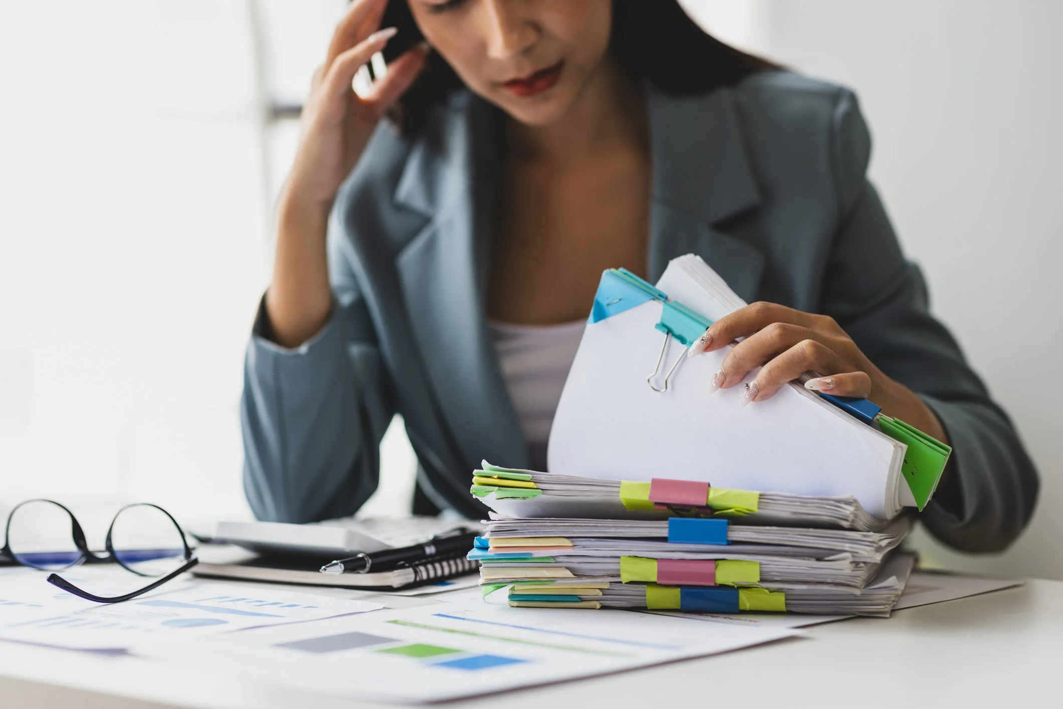 A woman sits at a desk, holding a stack of papers and folders with colorful tabs. She appears to be talking on the phone and is surrounded by organized files, a pen, and a pair of glasses on the desk.