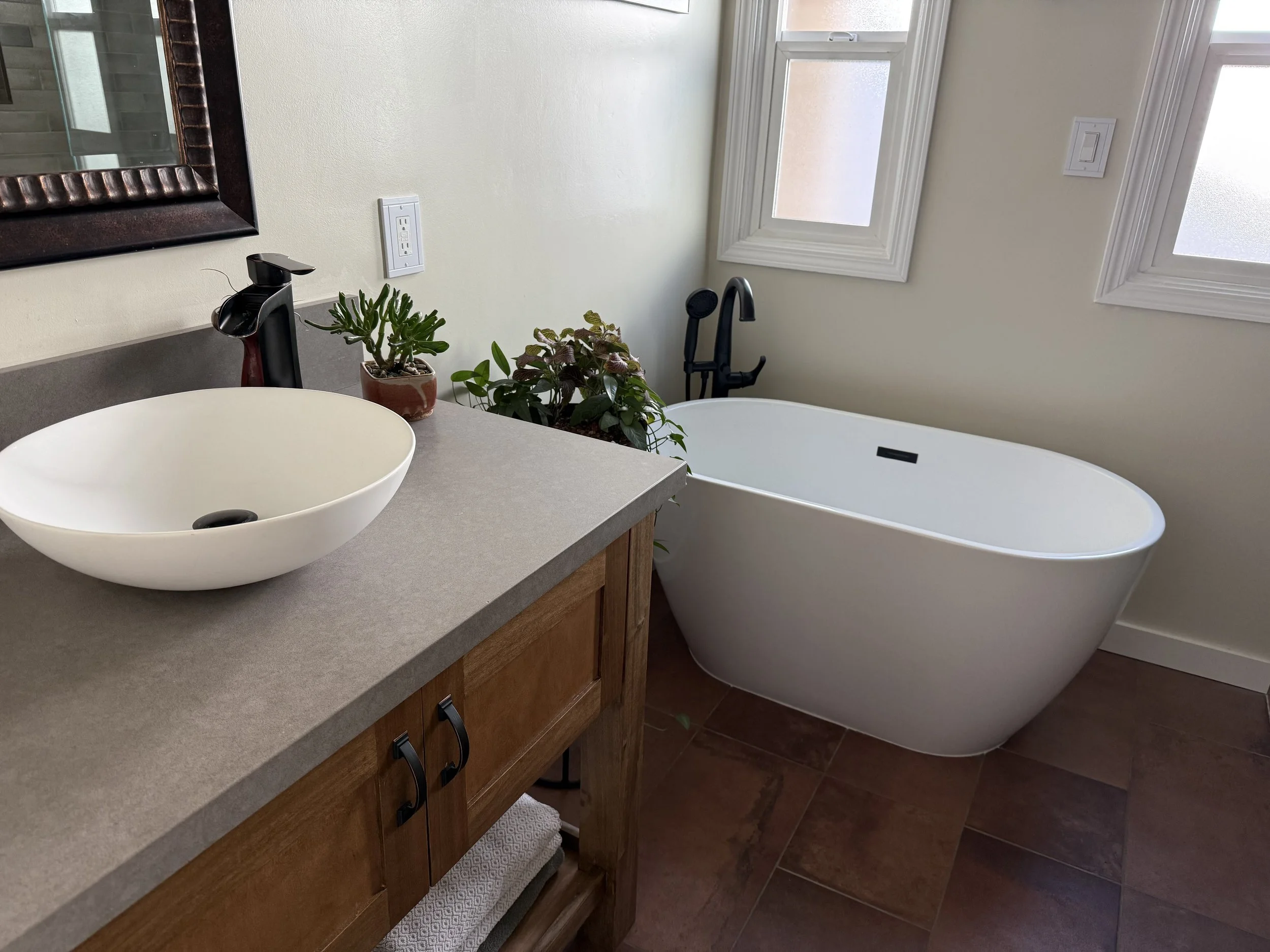 Bathroom with a white bathtub next to a window with frosted glass, a wooden cabinet with a white sink and black faucet, two potted plants on the counter, and a mirror on the wall.