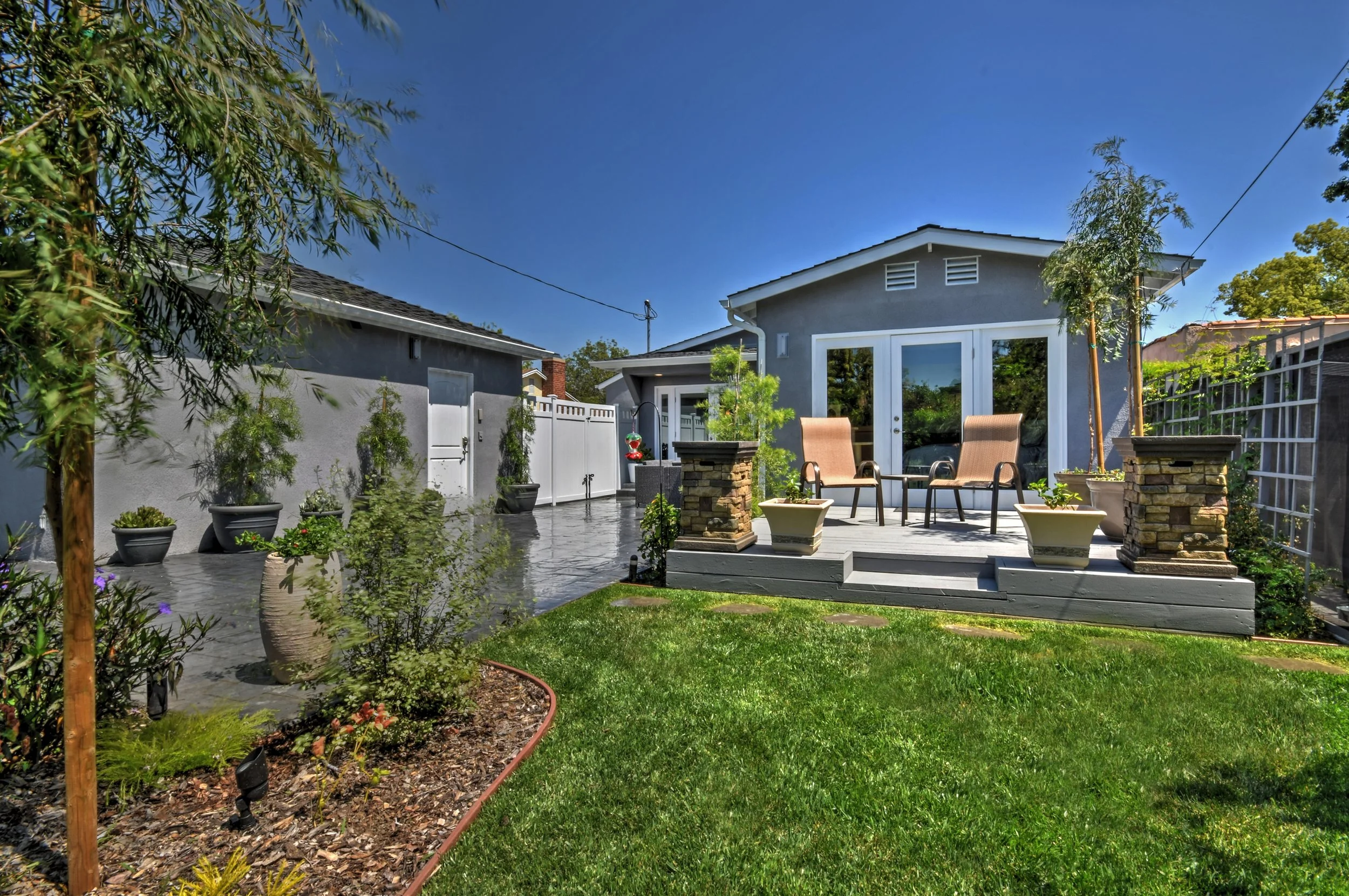 A backyard patio with two beige chairs surrounded by potted plants and flowers, a small tree, and a wooden deck with sliding glass doors leading into a gray house under a blue sky.