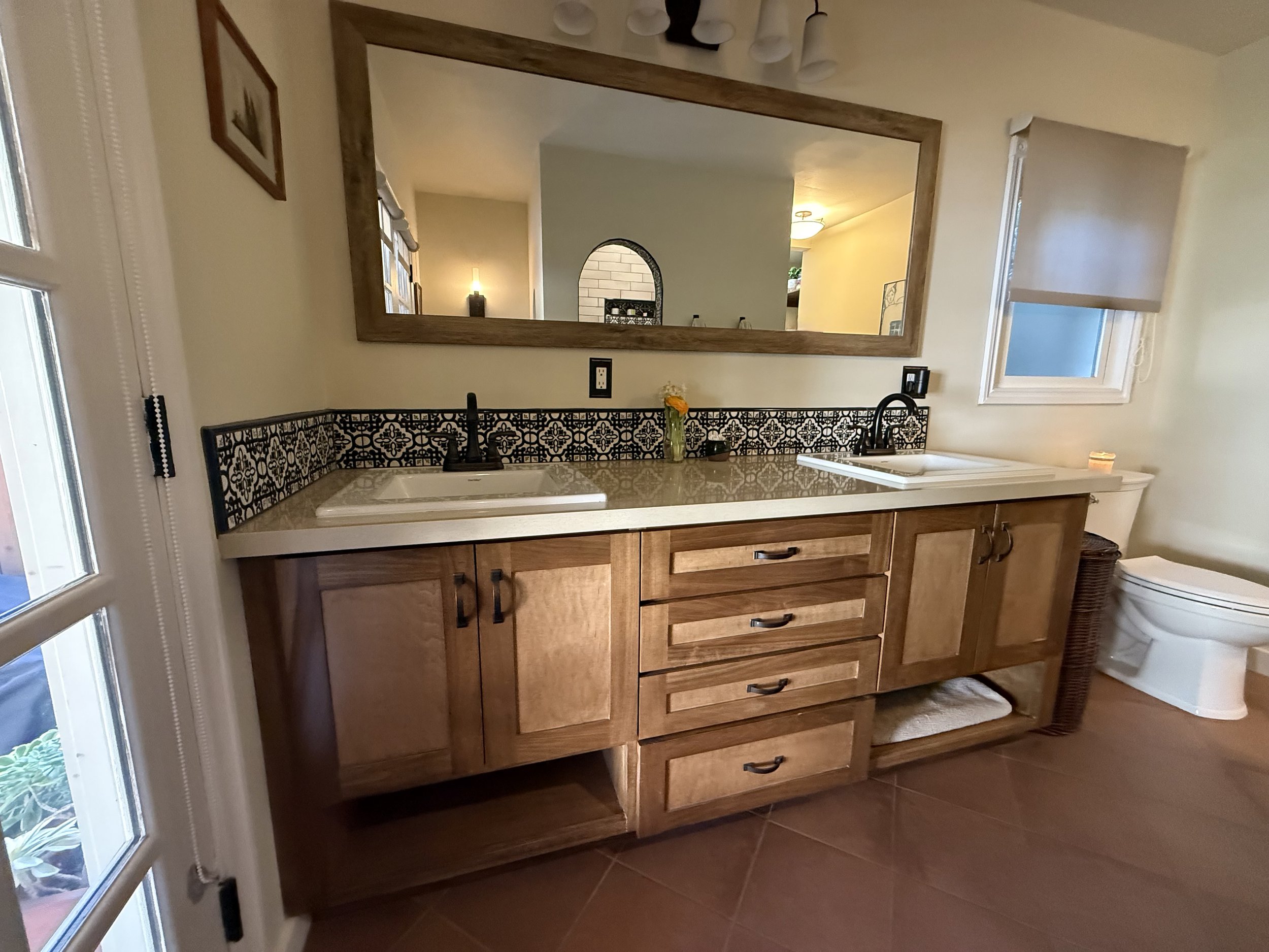 A kitchen with a wooden cabinet and drawers, a white sink, and a black decorative tile backsplash. There are two black light switches and a small yellow flower in a vase on the countertop. A large mirror hangs above the sink, reflecting part of the r