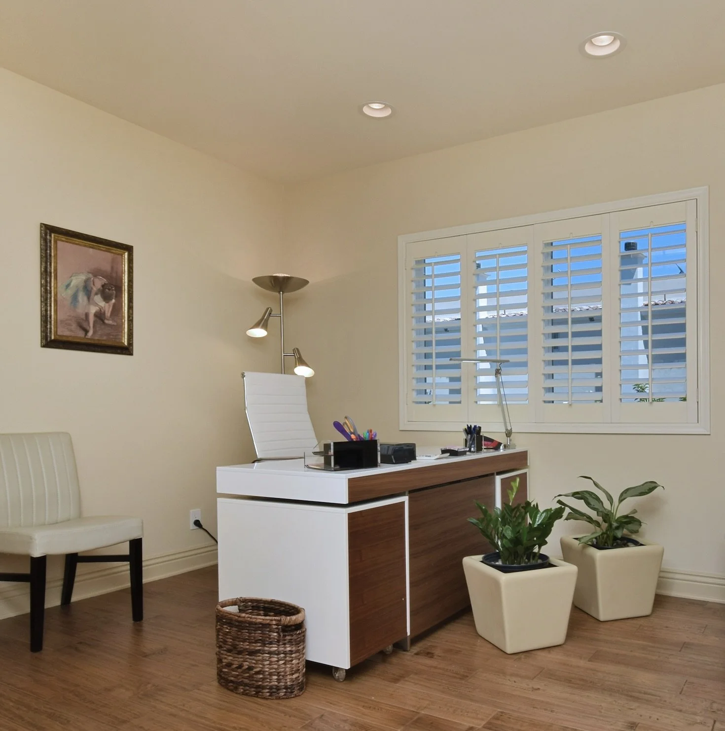 An office with a white desk, white chair, potted plants, wall art, and window blinds.