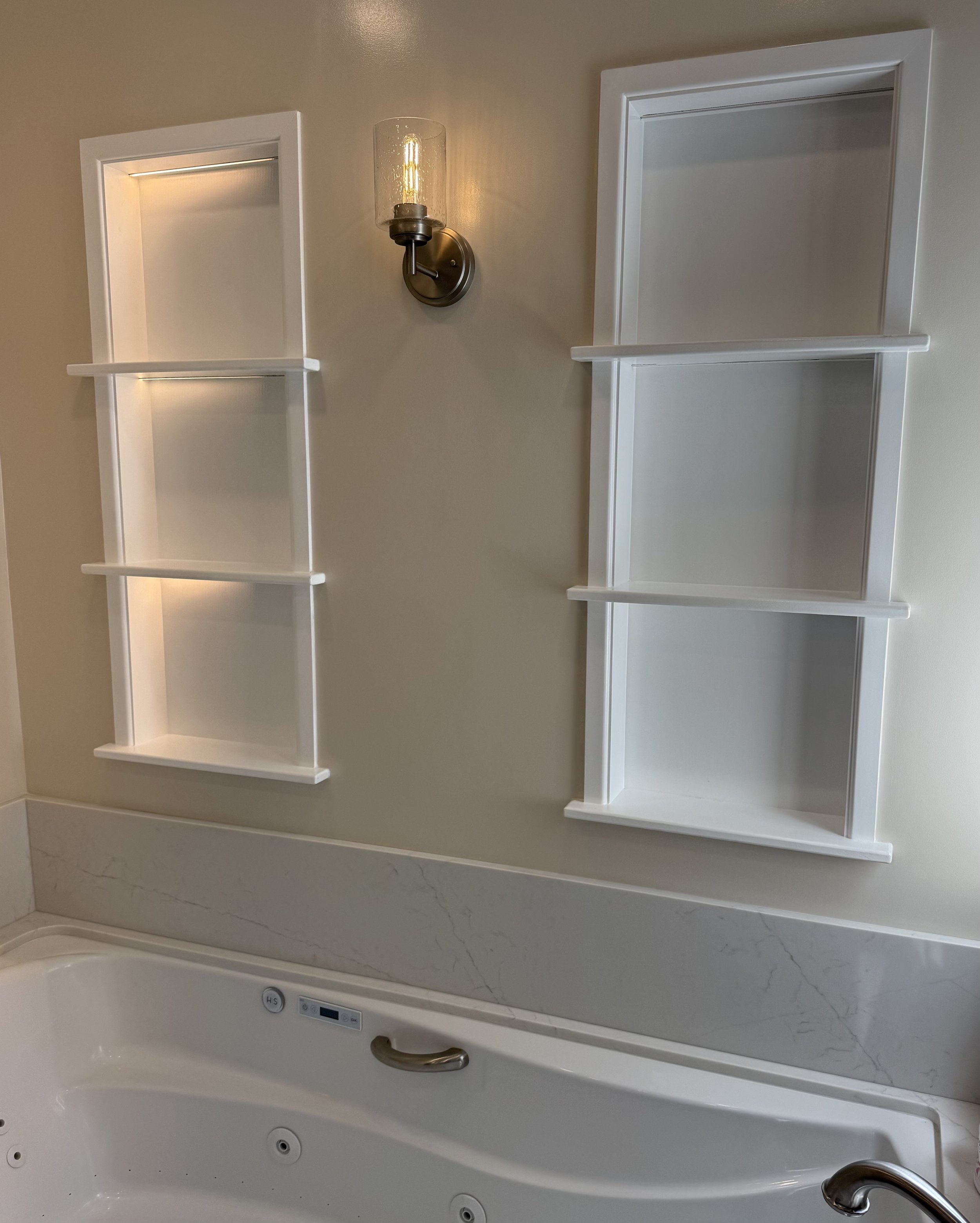 A bathroom with a large whirlpool bathtub with a chromed handle, white marble-like surround, and two white wall-mounted corner shelves, illuminated by a wall-mounted light fixture with exposed filament bulb.