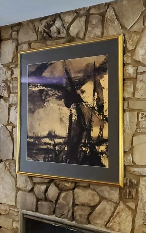 Abstract painting of a landscape featuring trees, a tower, and a sky, in dark and earthy tones, framed in gold and mounted on a stone wall.