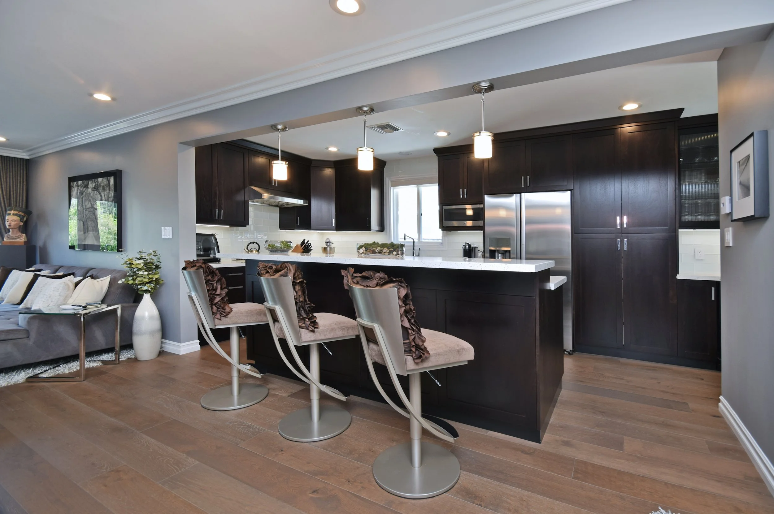 Modern kitchen with dark wood cabinets, white countertops, and stainless steel appliances, with a dining area featuring beige chairs with ruffled fabric and a row of pendant lights overhead.