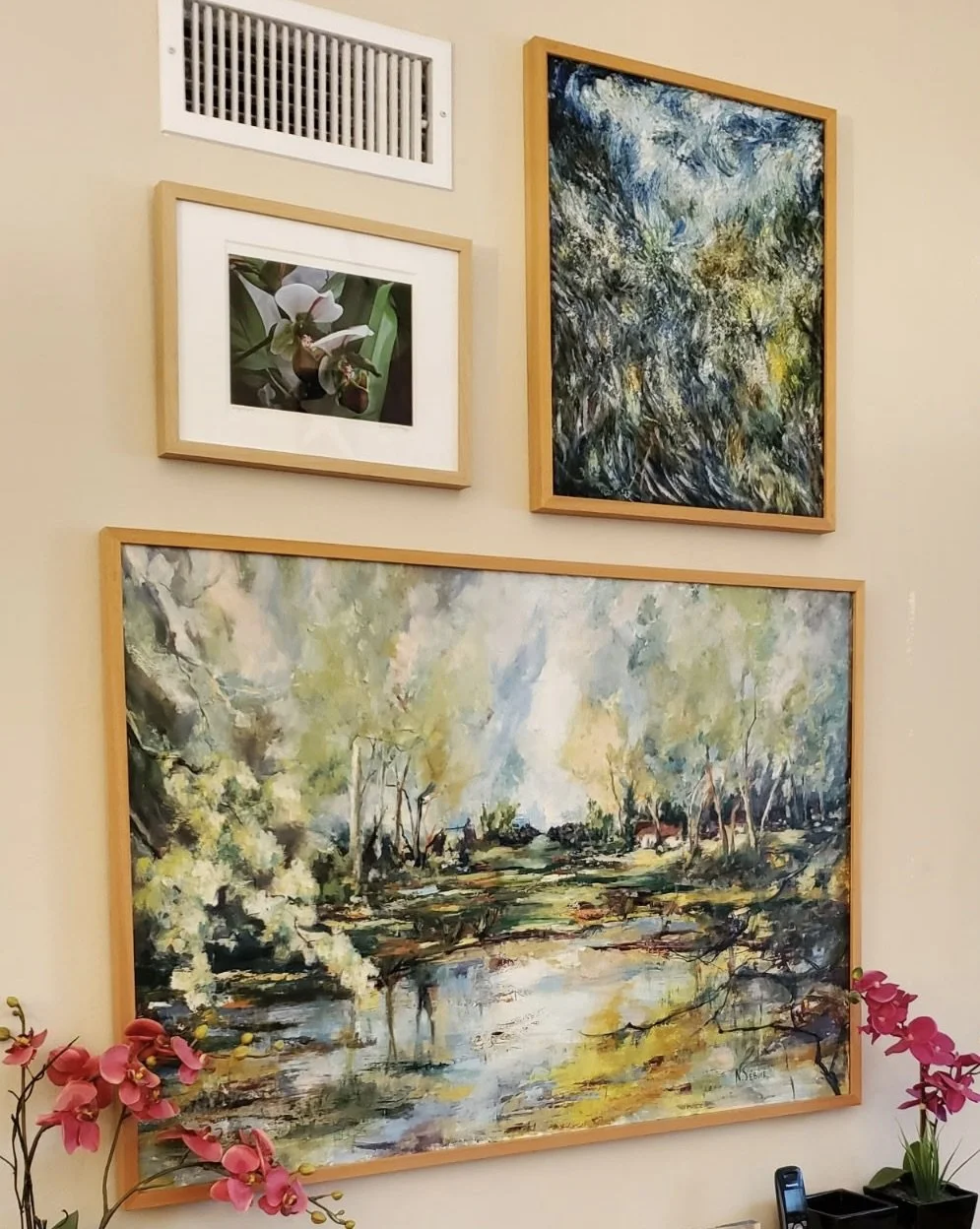 A wall decorated with four framed artworks including a photograph of orchids, two abstract paintings with green and blue tones, and a landscape painting of a creek with trees and reflections in water, with pink orchids at the bottom.