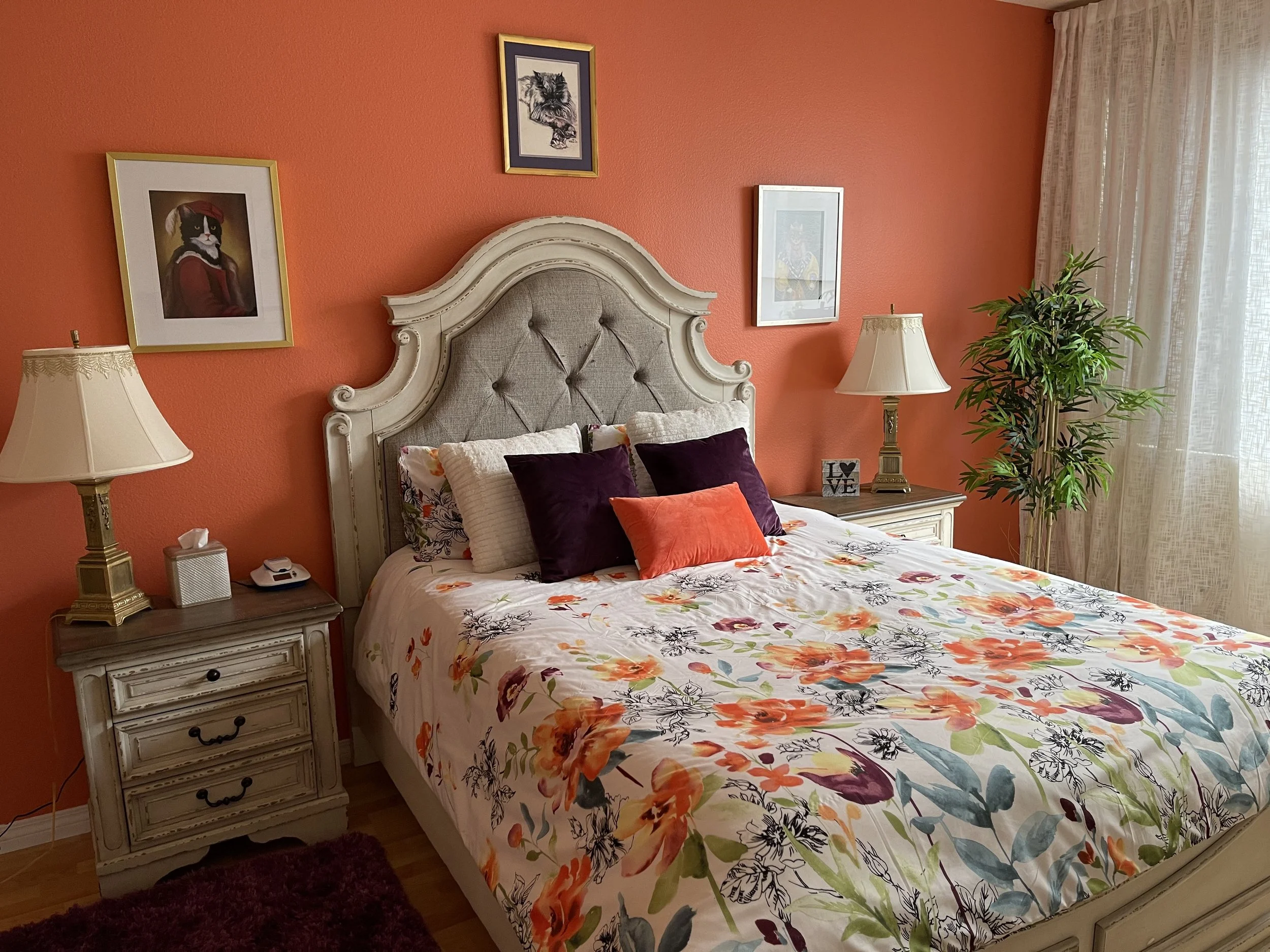 A bedroom with an orange wall, a white bed with floral bedding, a white headboard, matching white nightstands with lamps, framed art on the wall, a tall green plant near the window with sheer curtains.