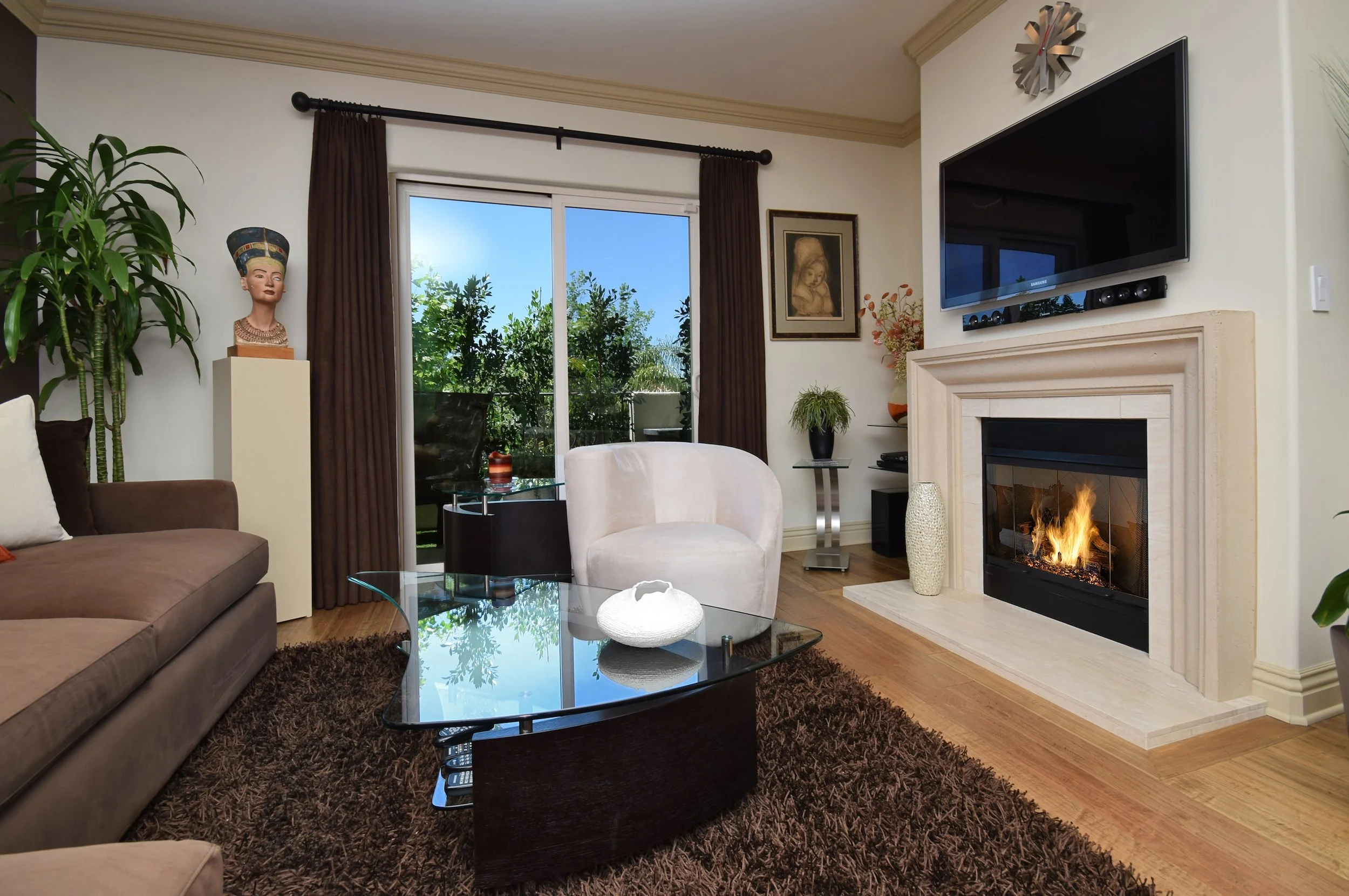 A living room with a brown sofa, a white armchair, glass coffee table, fireplace with a flame, large flat-screen TV, and window with brown curtains showing greenery outside.