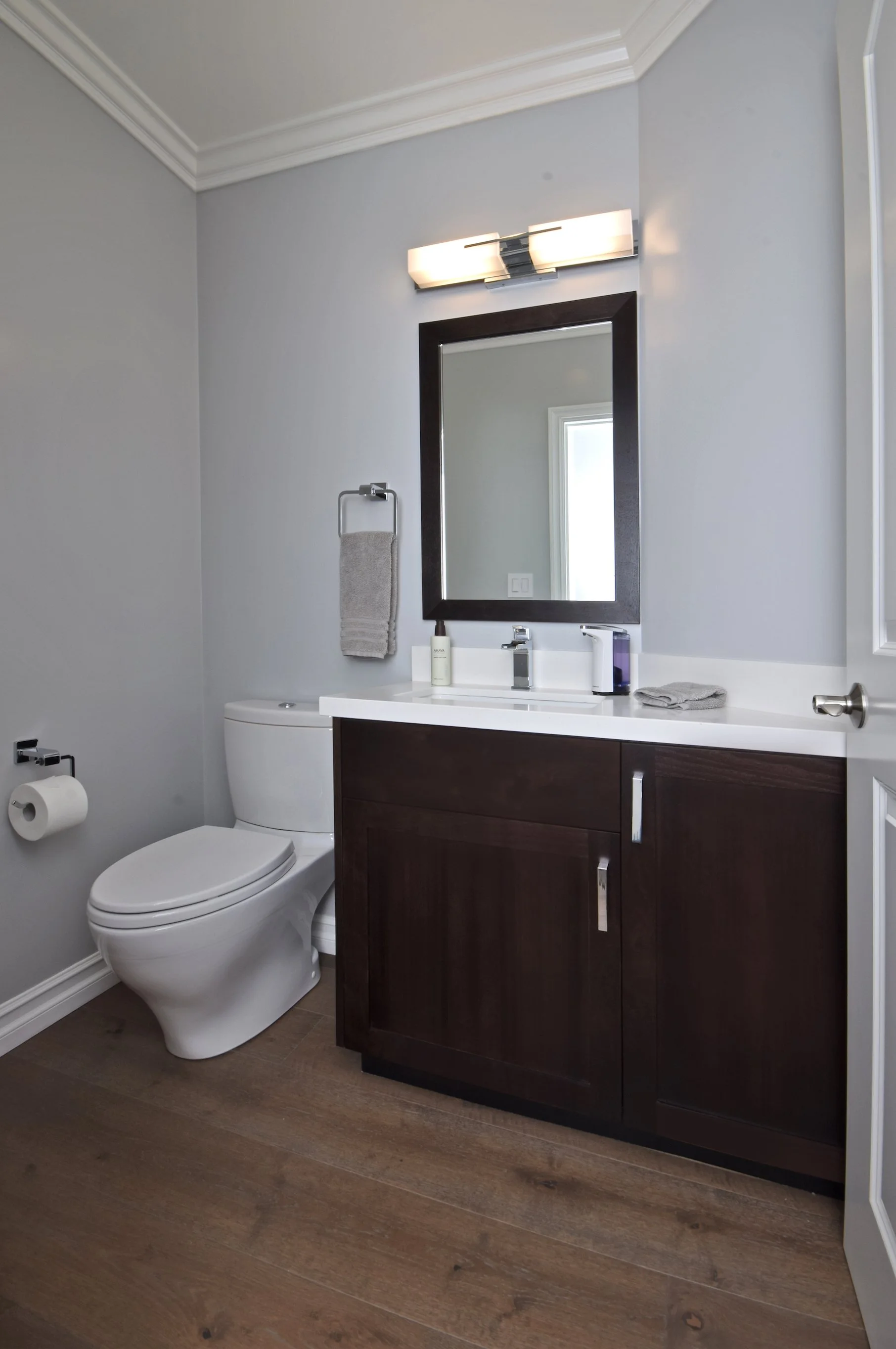 A modern bathroom featuring a white toilet, a dark wooden vanity with a white countertop, and a rectangular mirror. The bathroom has light gray walls, crown molding, and a wooden floor. A towel is hanging on a towel ring beside the mirror, and toilet