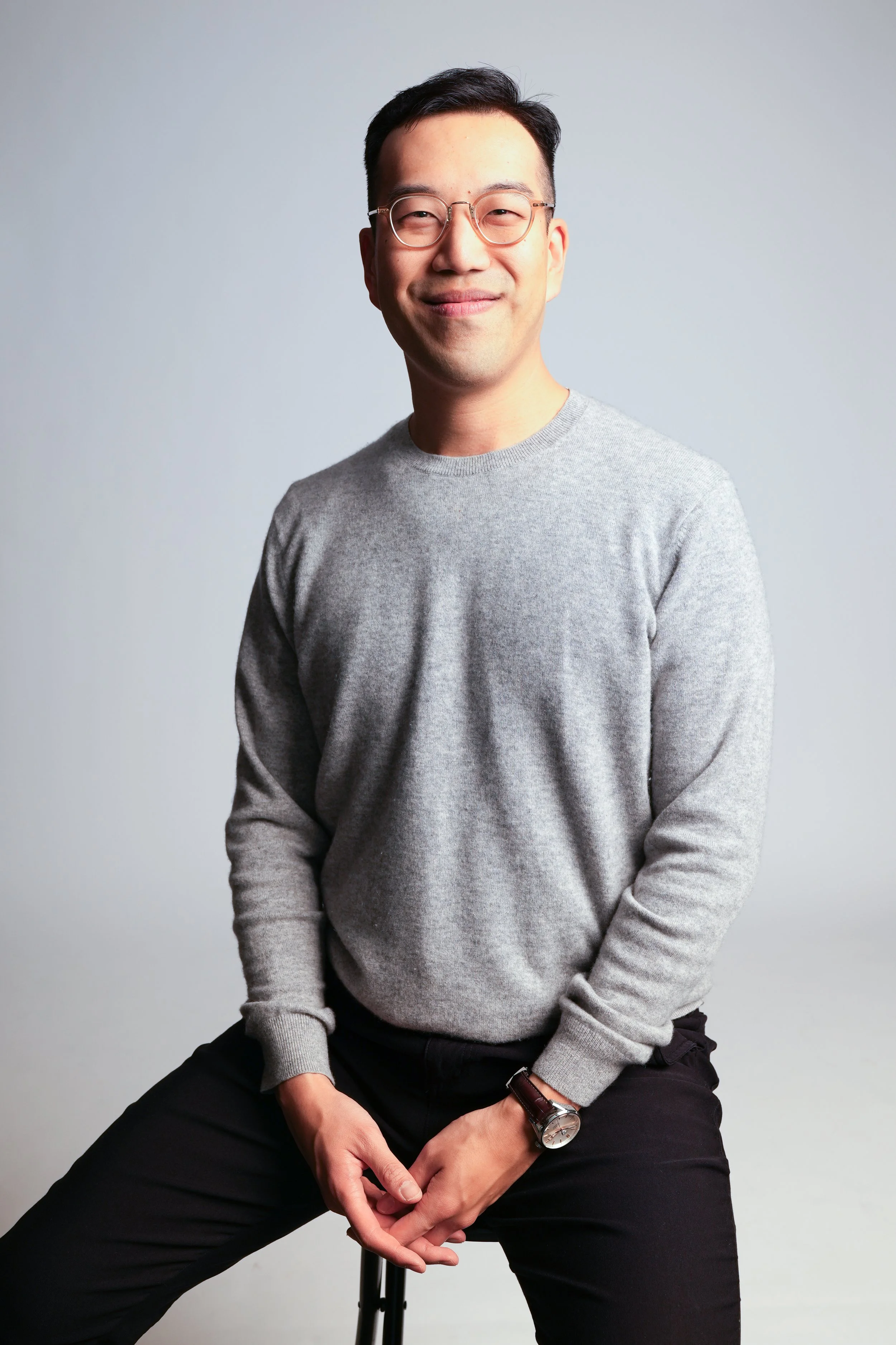 A man with glasses in a grey sweater sitting on a stool against a light grey background, smiling at the camera.