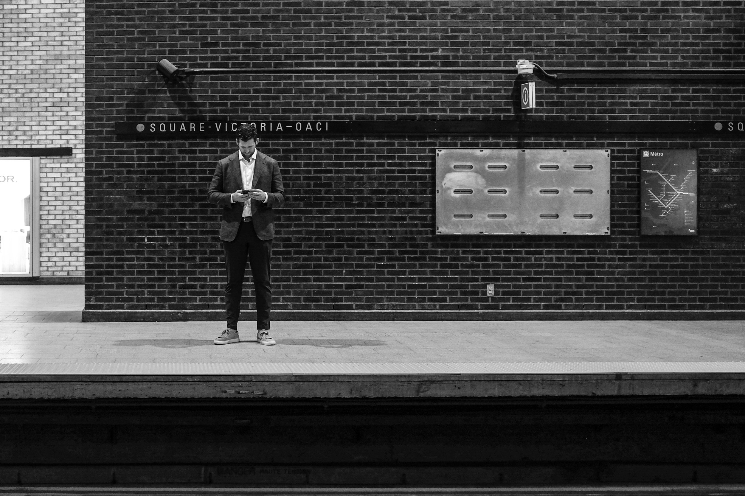 A man in a suit standing on a subway platform at Square-Victoria station in Montreal, looking at his phone, with a brick wall and subway signs in the background.
