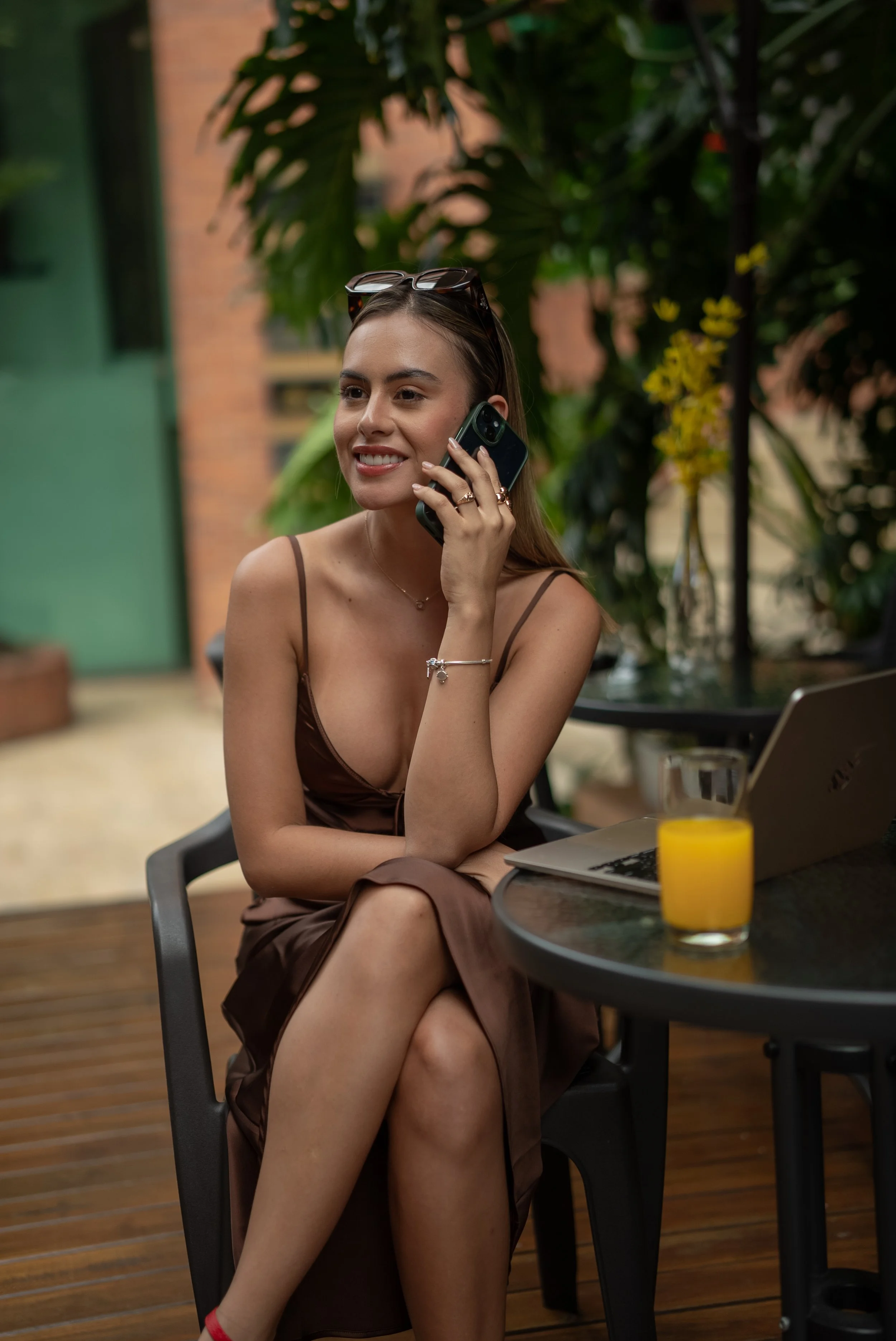 Una mujer sonriente con cabello castaño y pantalón largo de seda en un entorno con plantas verdes y un fondo de ladrillos, sentado en una mesa de madera, hablando por teléfono móvil y frente a una computadora portátil, con una vela amarilla en la mes