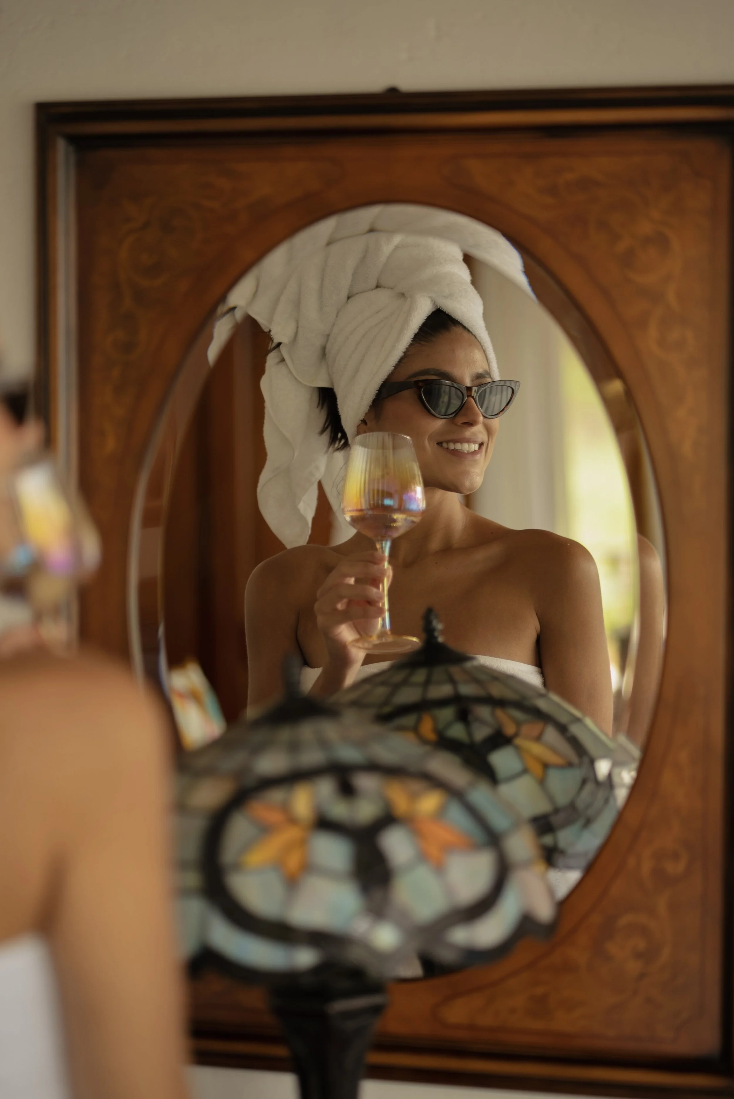 Mujer con toalla en la cabeza y gafas de sol, sonriendo y sosteniendo una copa con bebida, reflejada en un espejo de madera con diseño.