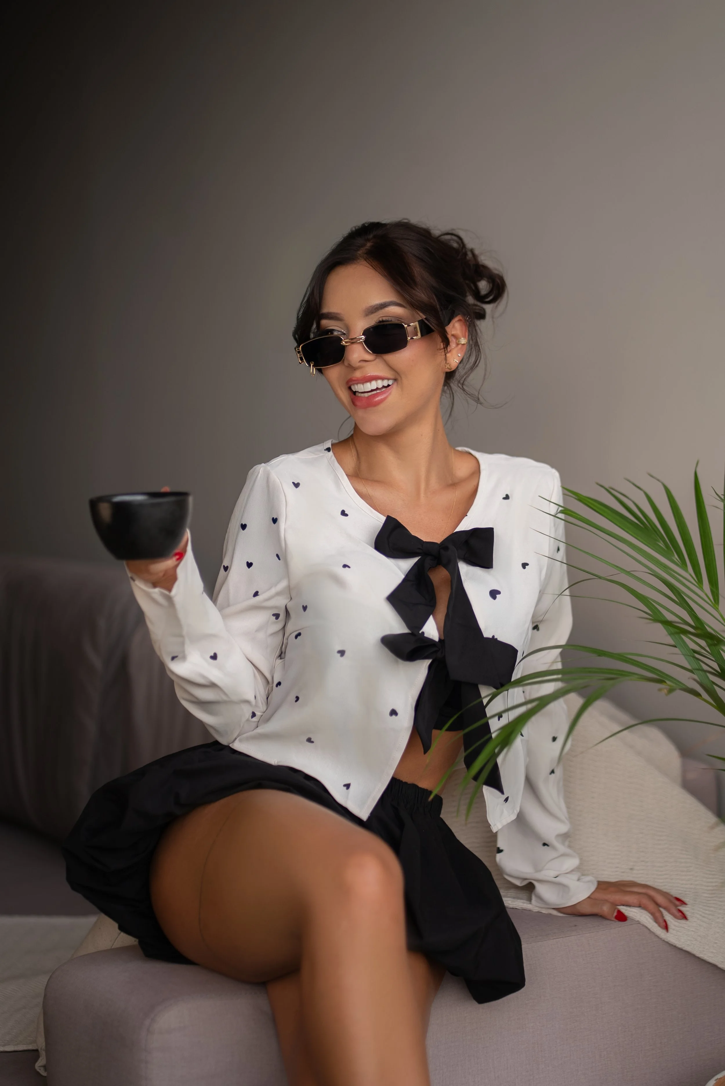 Mujer joven con gafas oscuras, camisa blanca con pequeños corazones negros y lazo negro, sosteniendo una taza, sentada en un sillón de sala, con plantas verdes al frente y fondo gris.