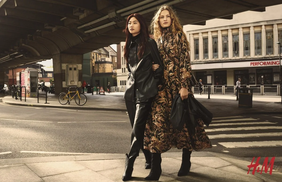 Dos mujeres de pie en la esquina de la calle, una con chaqueta de cuero negra y la otra con vestido largo con patrón floral, ambas con botas negras, en un entorno urbano con edificios y un puente sobre la calle, logotipo de H&M en la esquina inferior