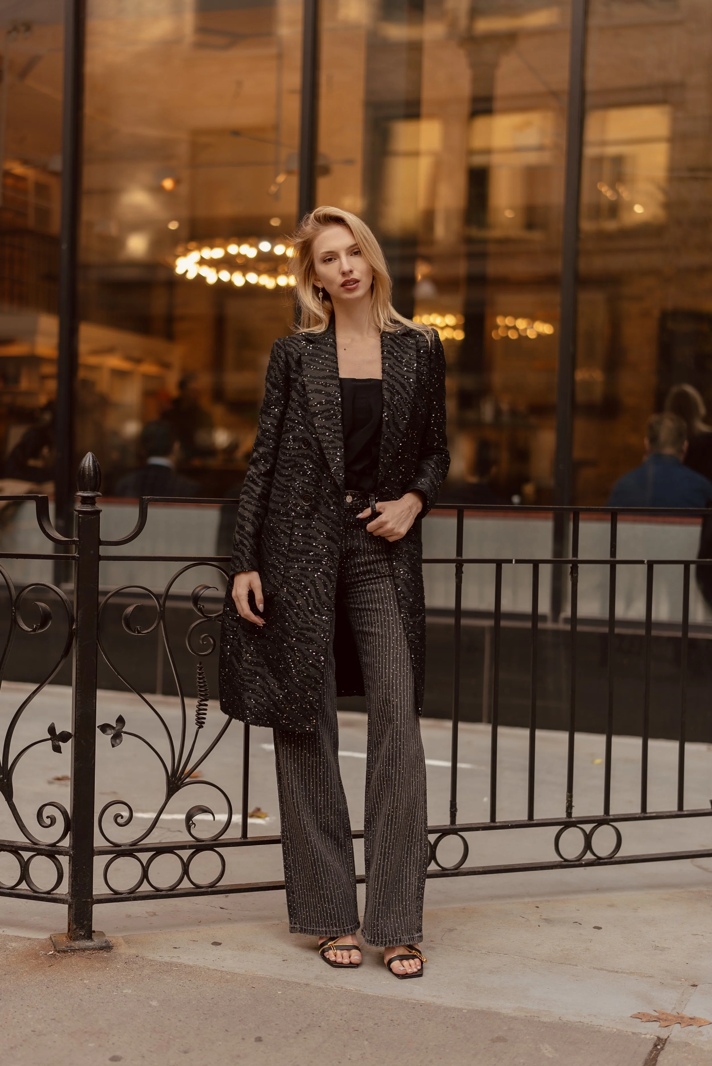 Mujer con pantalones de vestir a rayas y blusa negra, con un blazer negro con brillo, de pie junto a una cerca de hierro negro, en un entorno urbano con fondo de vidrio y luces cálidas en la noche.