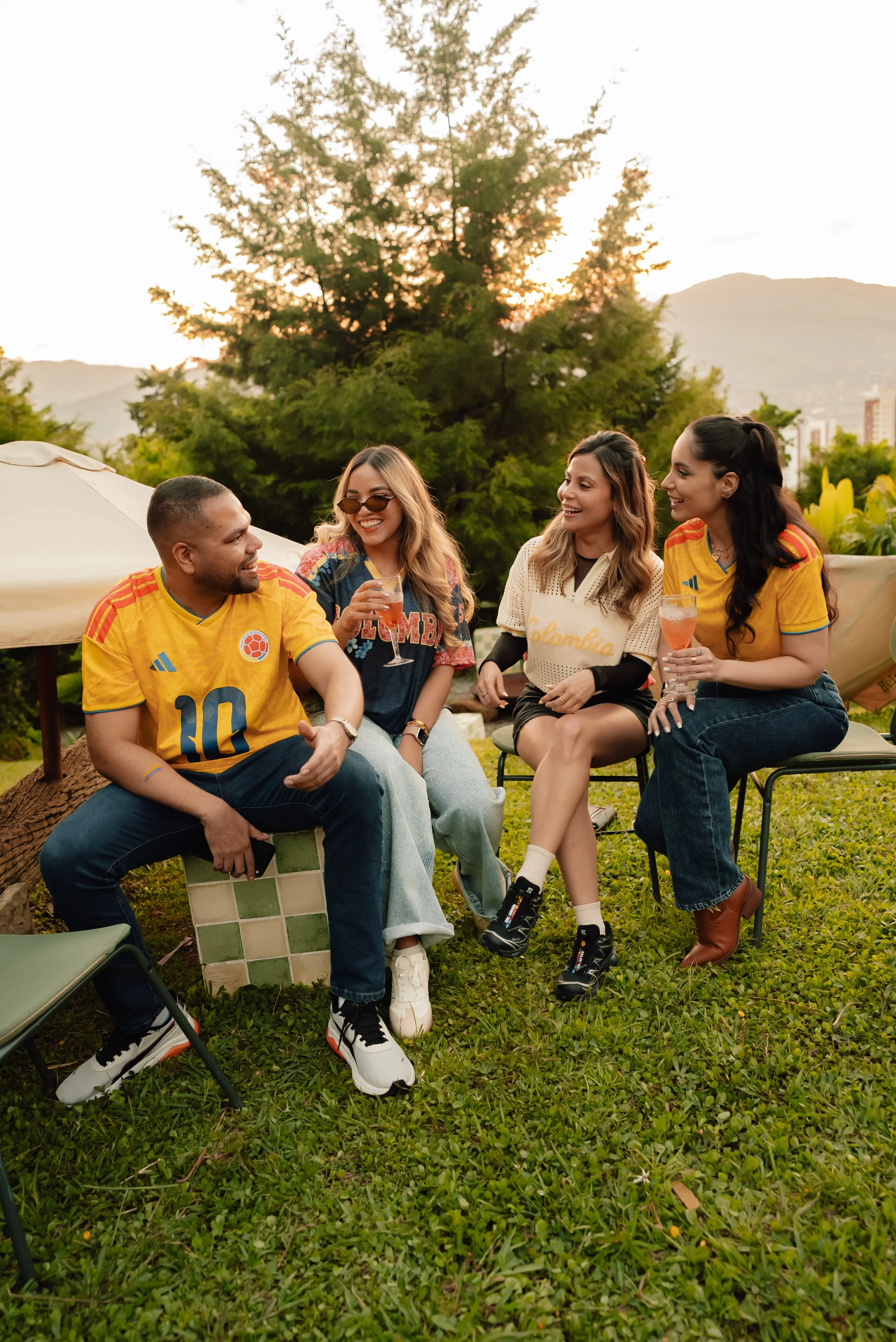 Grupo de cinco personas jóvenes sentadas y conversando en un jardín durante el atardecer, con árboles y montañas de fondo, algunas sosteniendo bebidas.