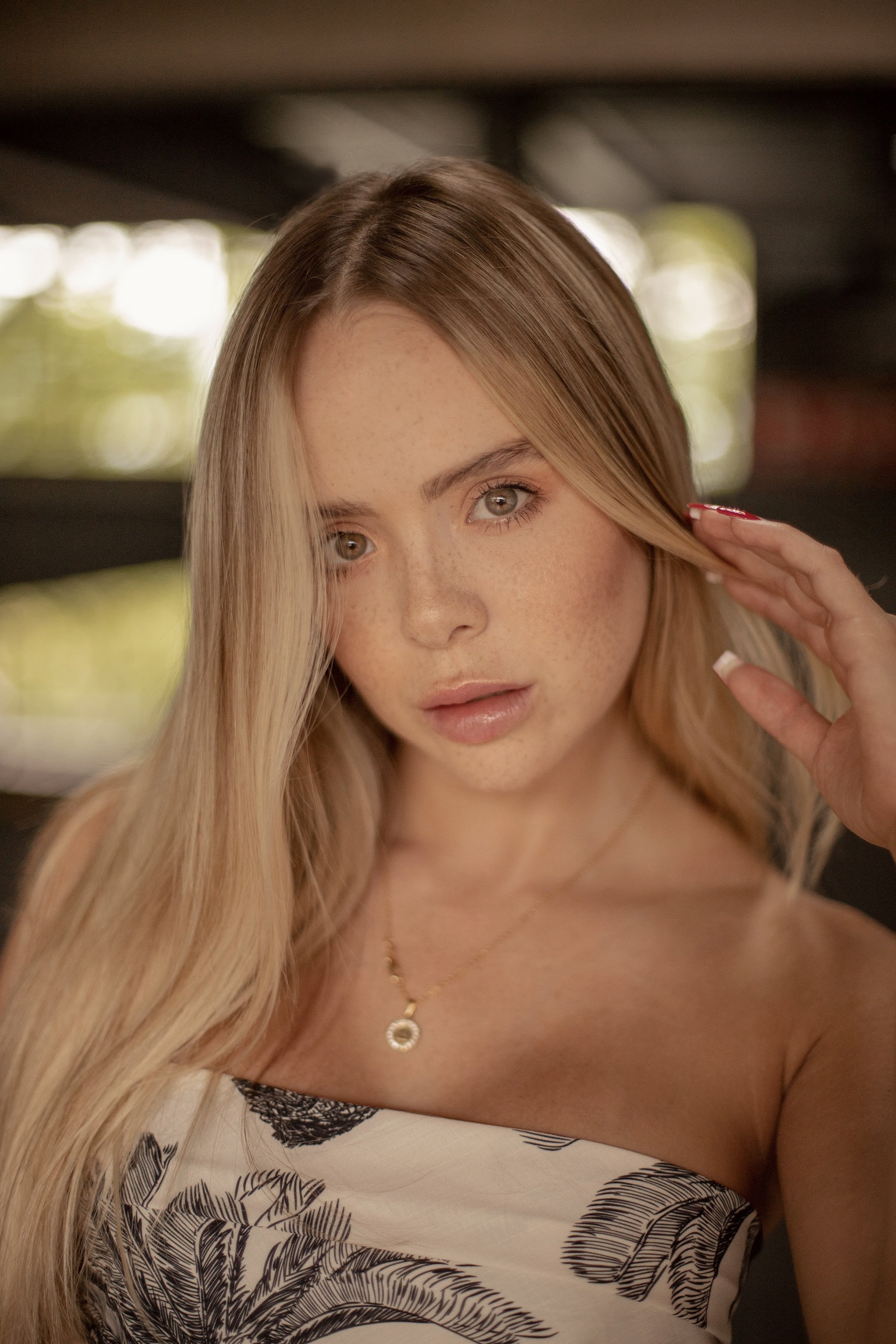 Primer plano de una joven con cabello rubio y ojos claros, tocándose el cabello con la mano, con fondo difuso y luces brillantes.