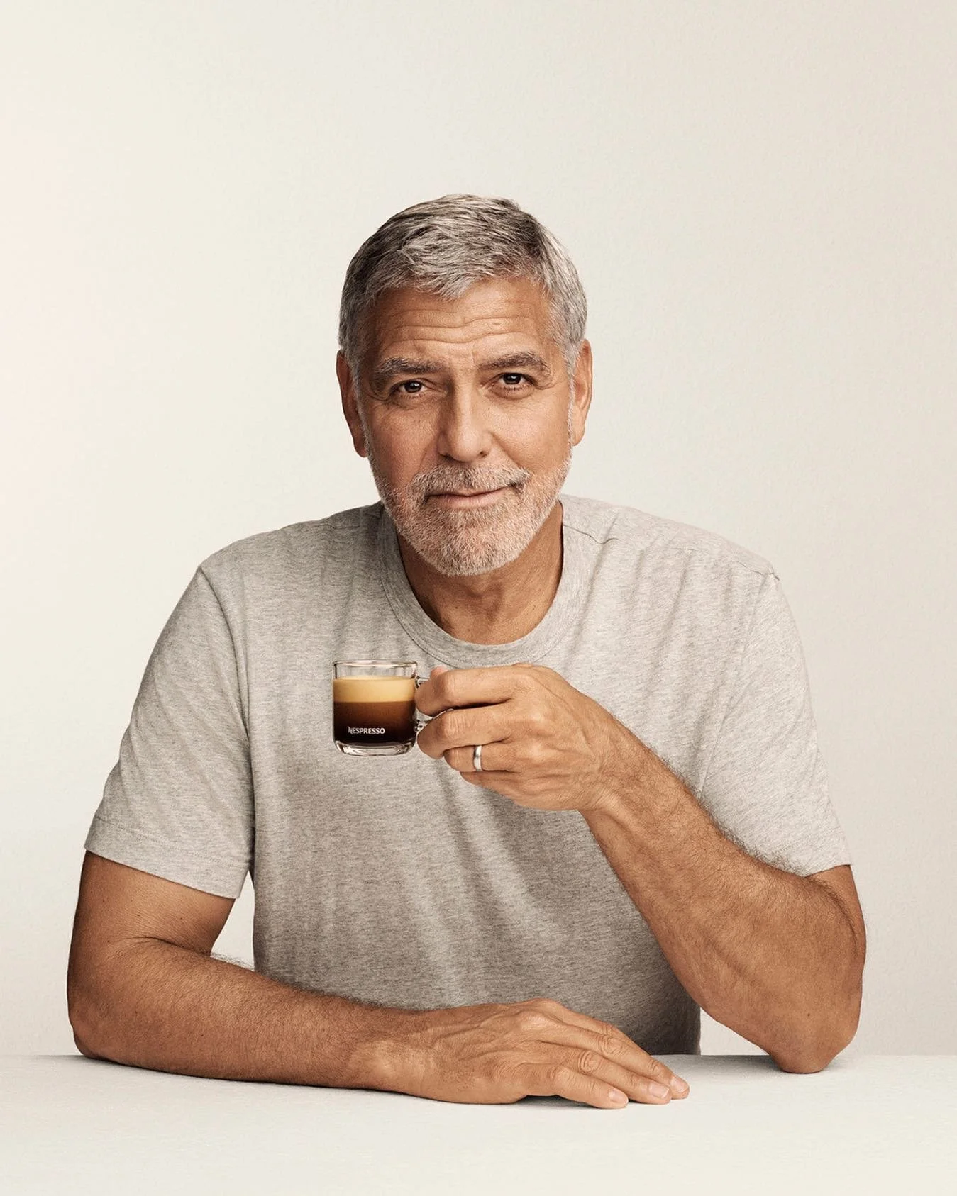 Hombre de cabello canoso y barba, con una expresión amable, sostiene una taza de café espresso en su mano derecha. Él está sentado con sus brazos sobre una mesa y lleva una camiseta gris.