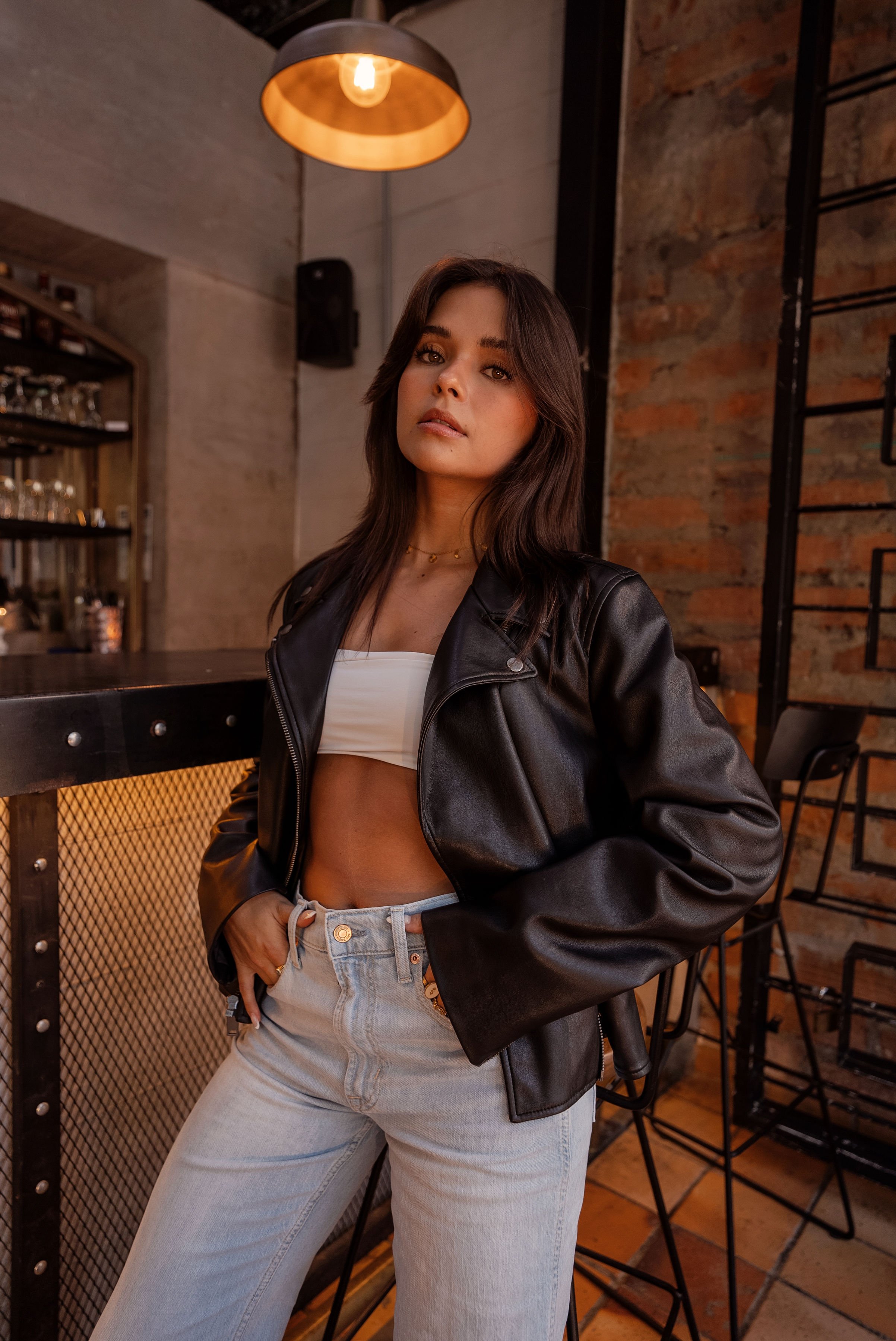 Mujer con cabello largo y oscuro, usando chaqueta de cuero negro y pantalones claros, en un ambiente de bar o restaurante con paredes de ladrillo y una lámpara de estilo industrial