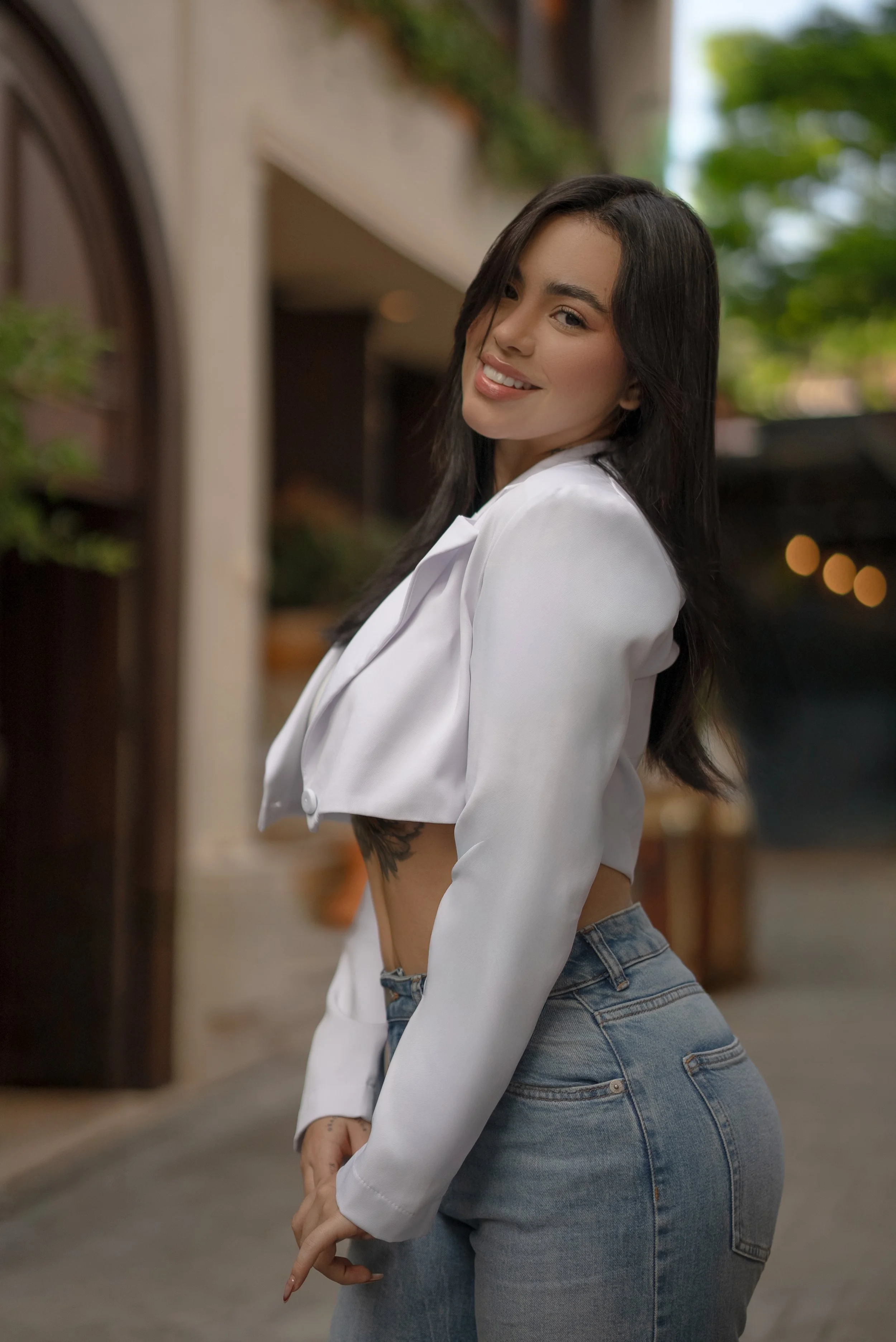 Joven mujer con blusa blanca y jeans, sonriendo, en un entorno urbano con paredes y plantas verdes.