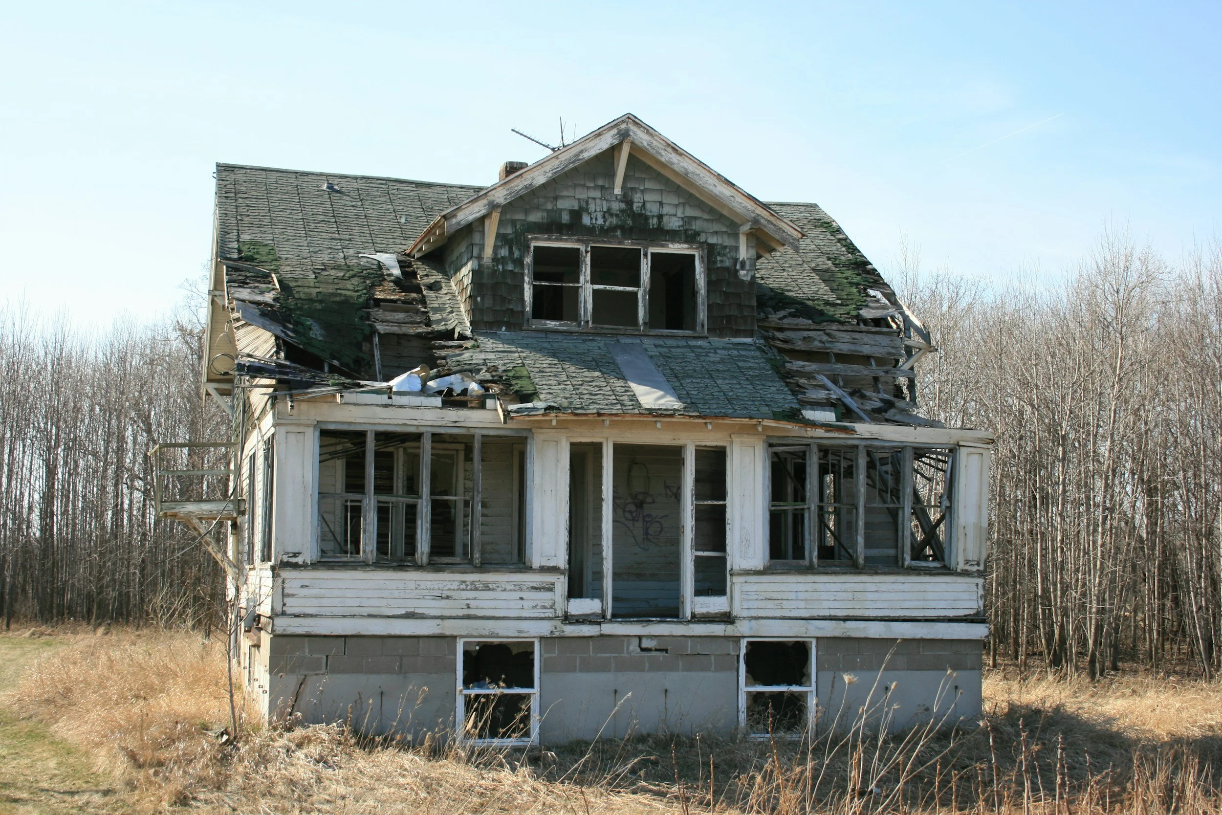 An abandoned, damaged two-story house with missing roof shingles, broken windows, and overgrown grass and weeds in front.