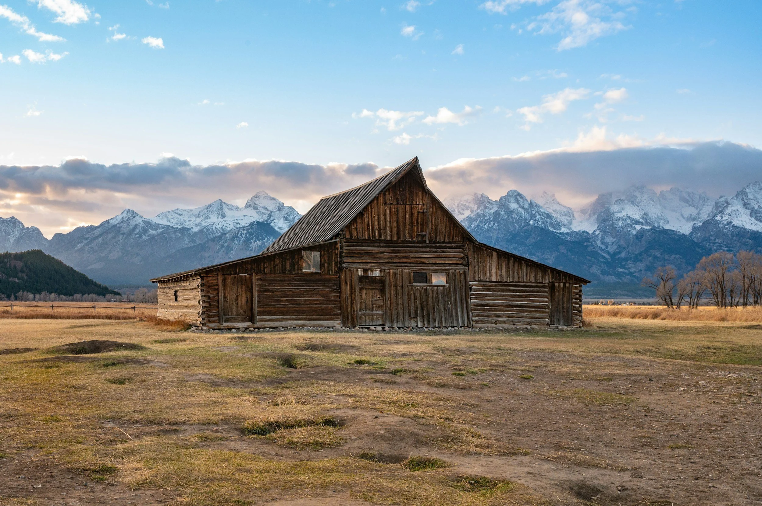 An old wooden barn in a field with mountains in the background, under a partly cloudy sky.