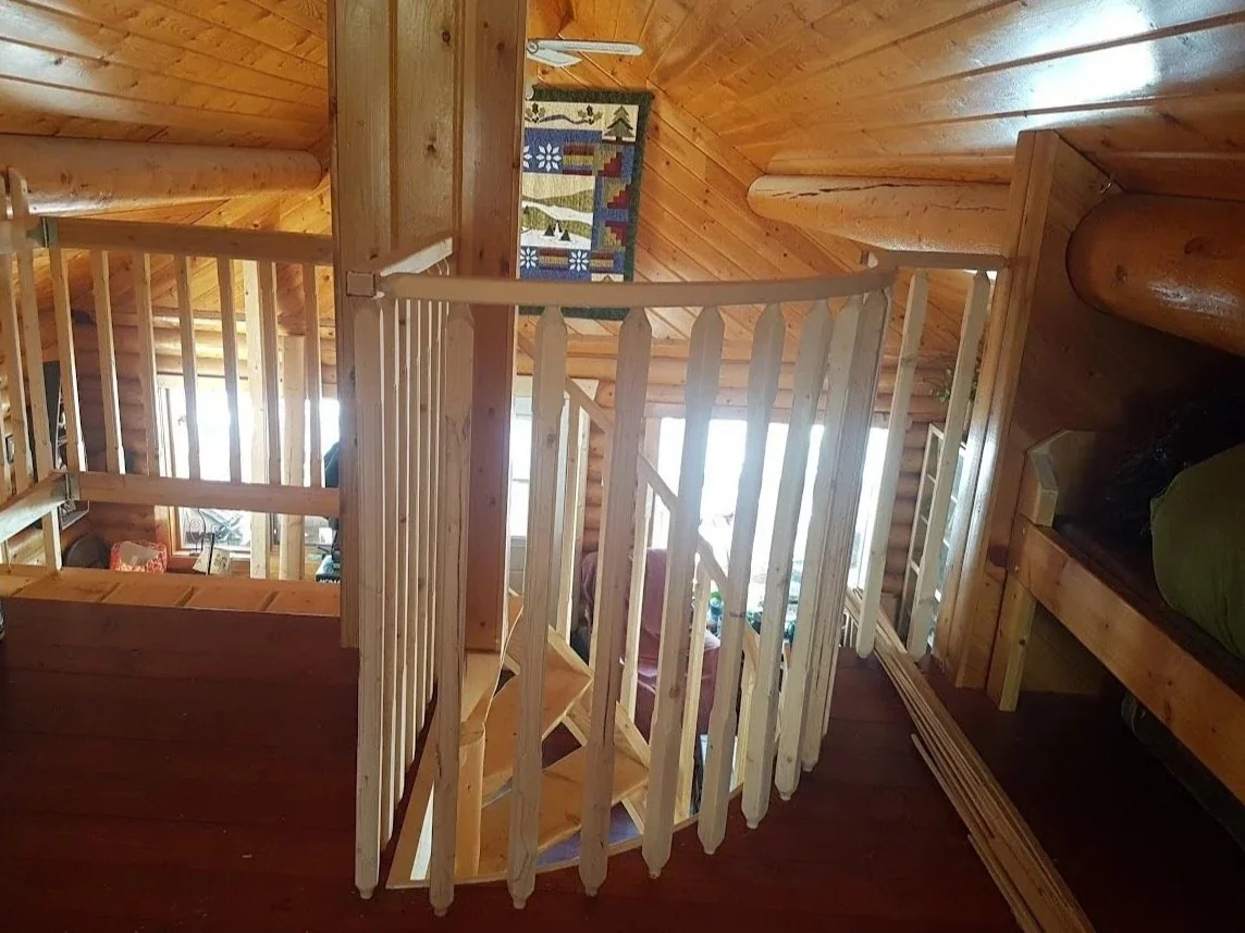 Interior view of a wooden spiral stairway in a log cabin, featuring a curved safety railing.