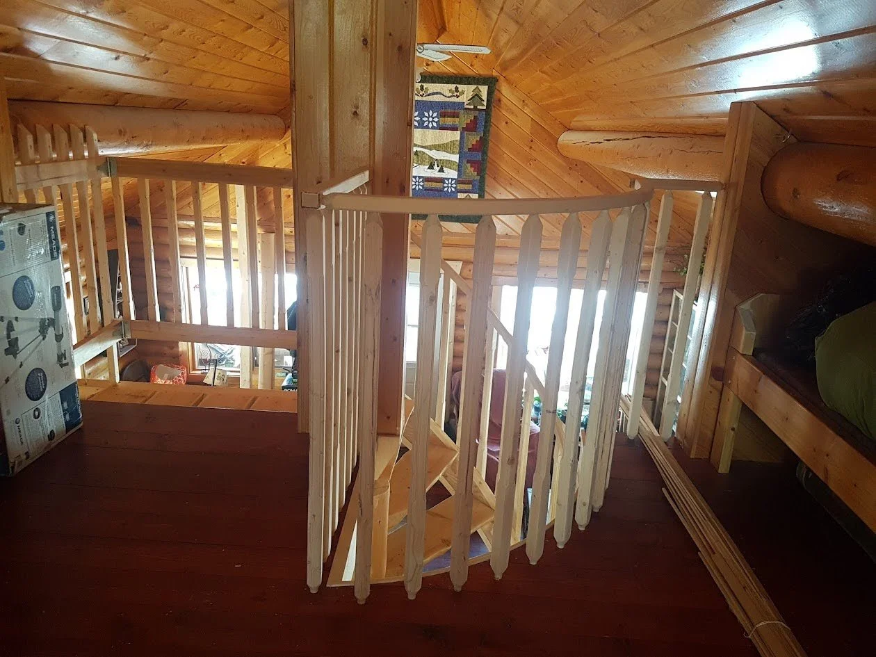 Interior view of a wooden spiral stairway in a log cabin, featuring a curved safety railing.
