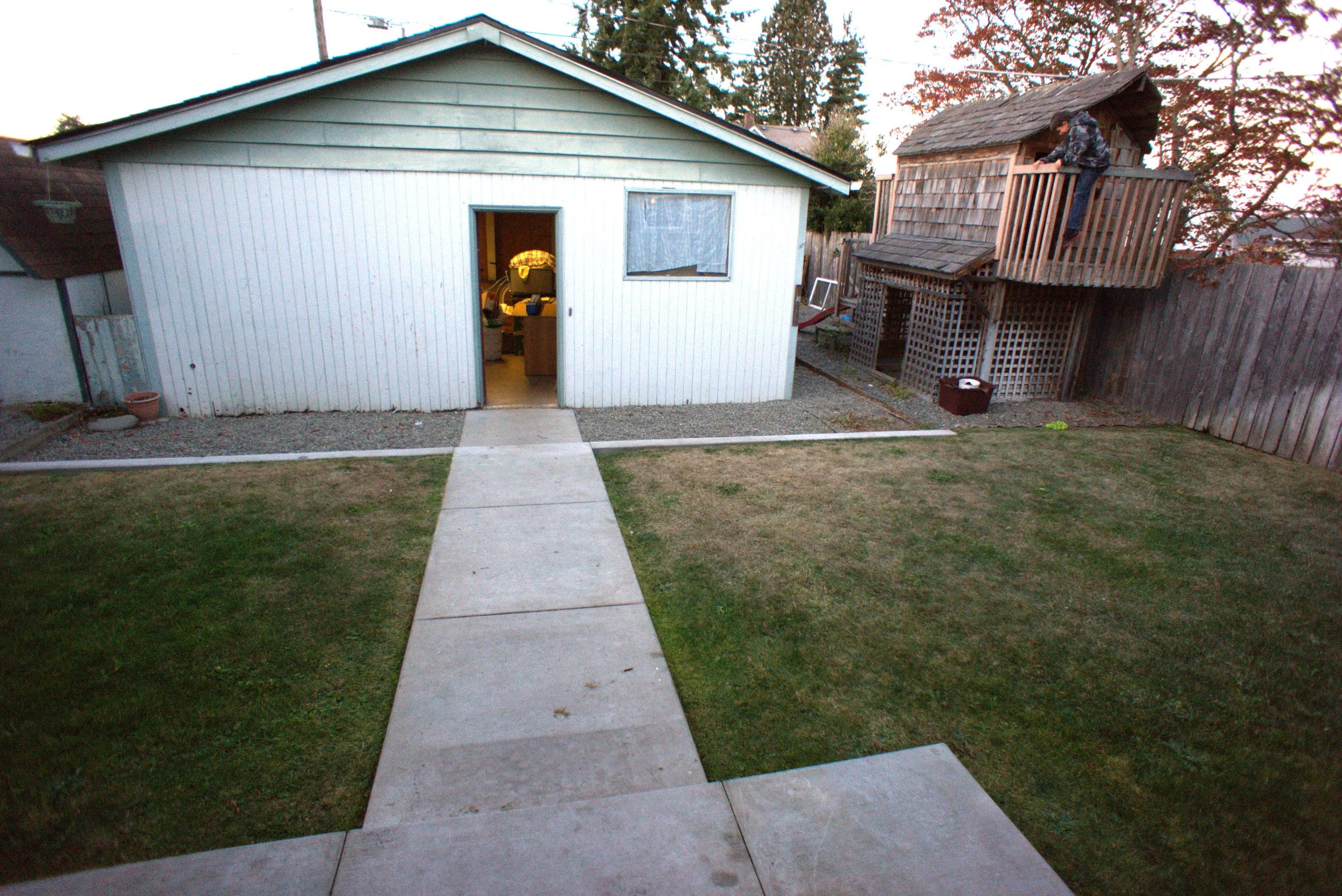 A backyard with a white garage, a concrete pathway, and a grassy lawn. There is a wooden playhouse to the right.