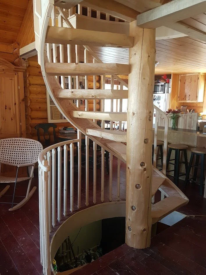 Spiral staircase made of light-colored wood in a log cabin interior with visible wooden walls and ceiling, leading to an upper level.