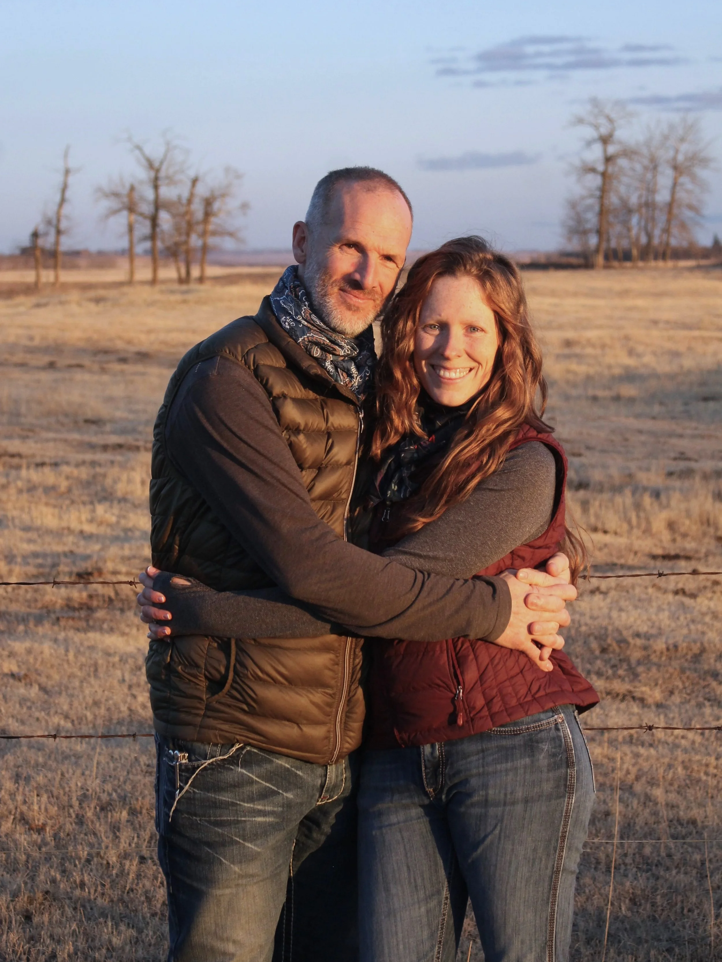 A man and woman hugging outdoors in a field with bare trees in the background during sunset