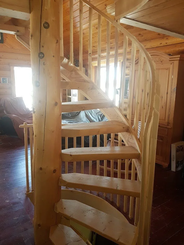 Wooden spiral staircase inside a cabin with natural wood paneling and furniture, sunlight coming through windows.