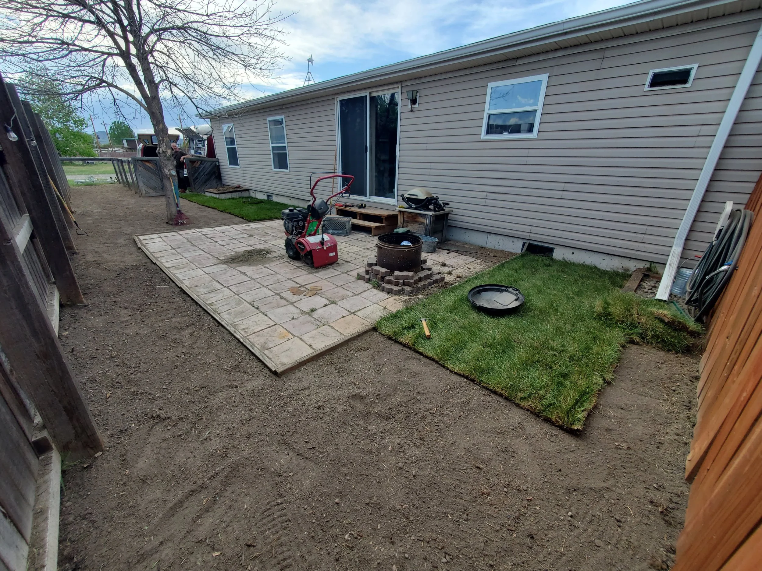 A backyard patio area with a small patch of freshly laid sod, a stack of pavers, a red rototiller, and various tools.