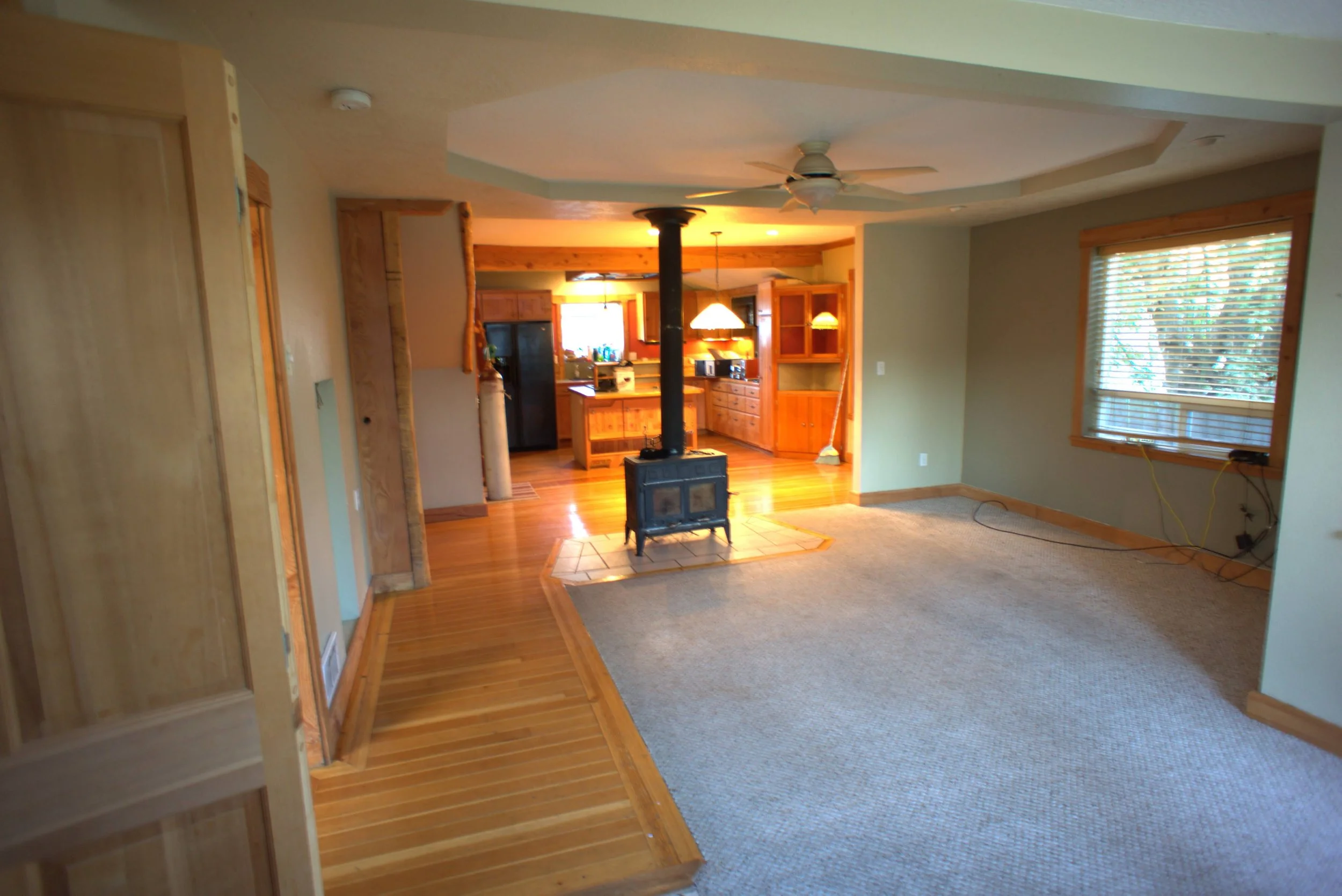 Interior view of a living room with a wood-burning stove, wood and carpet flooring, and an open kitchen area in the background with wooden cabinets.
