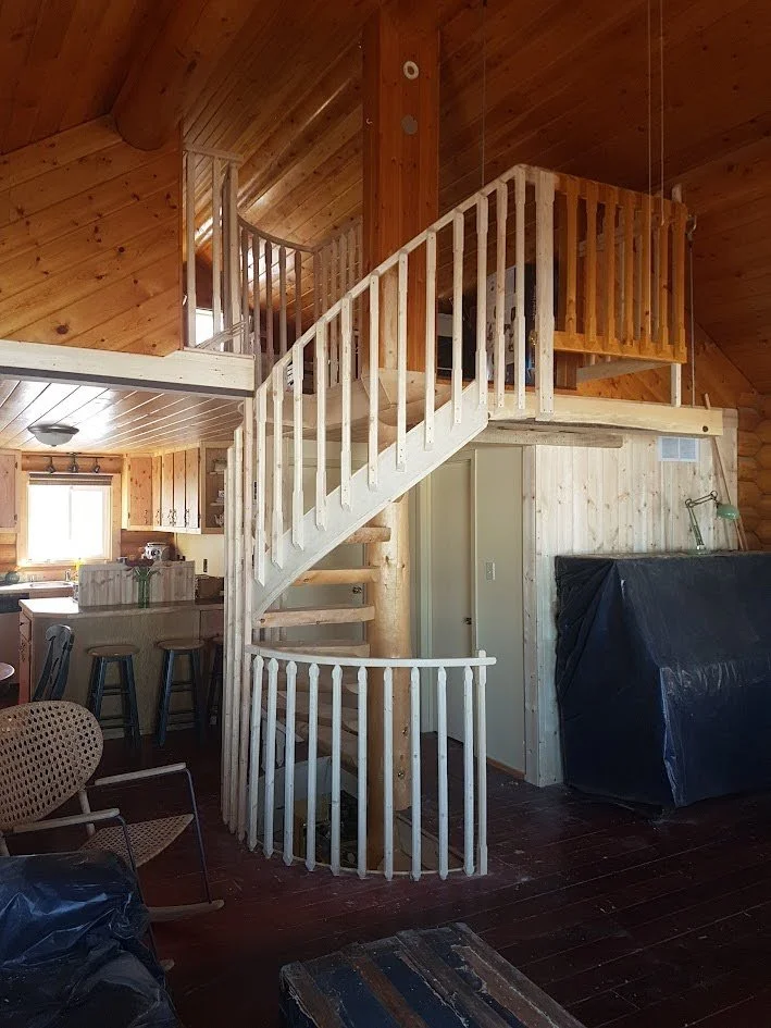 Interior of a log cabin with a spiral staircase made of wood leading to a second floor balcony.