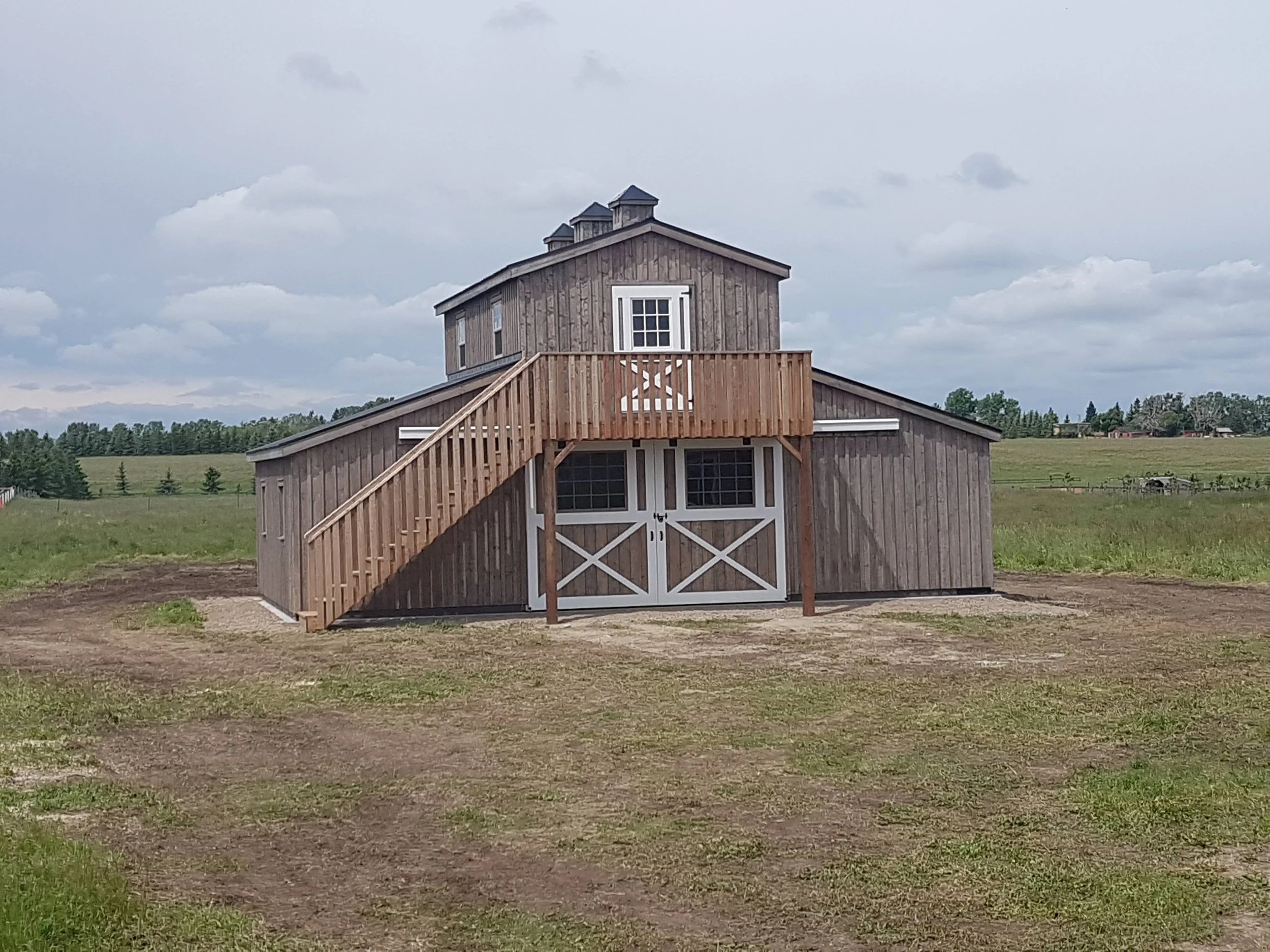 A large wooden barn with a rear balcony and stairs set in a grassy field under a cloudy sky.
