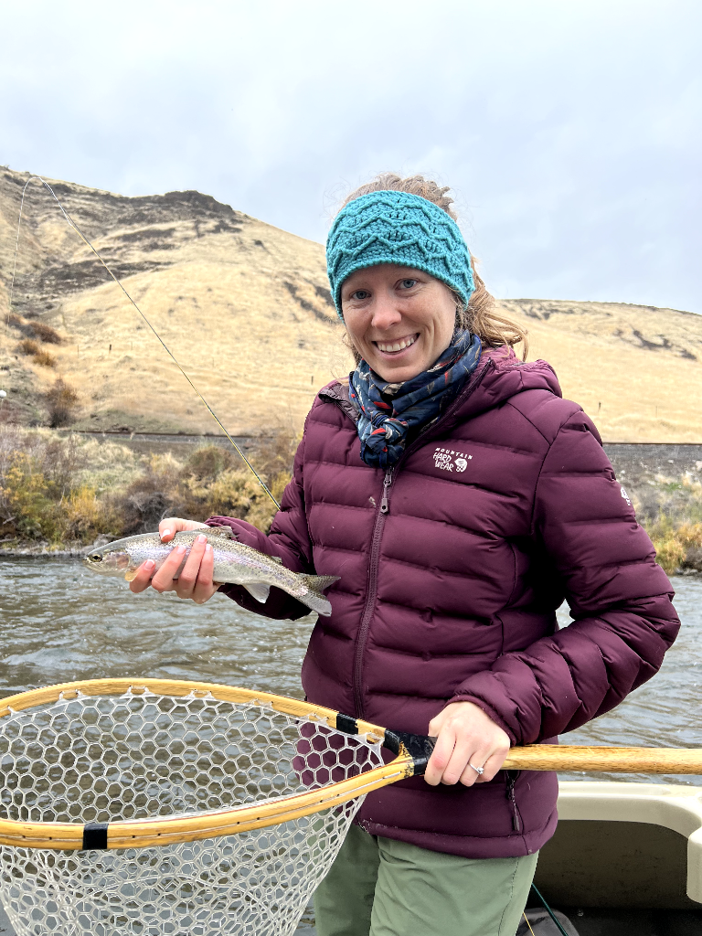 A woman in a purple jacket and teal knit headband holding a freshly caught fish on a fishing trip in a river, with hills in the background.