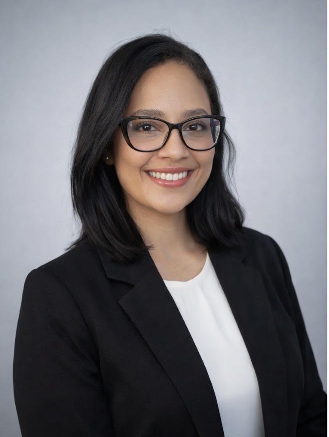 Professional woman with shoulder-length black hair, wearing glasses, a black blazer, and a white blouse, smiling against a plain light gray background.