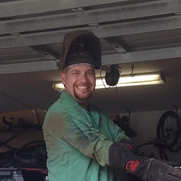 A man wearing a welding helmet on his head and a green work shirt, smiling in a garage or workshop.