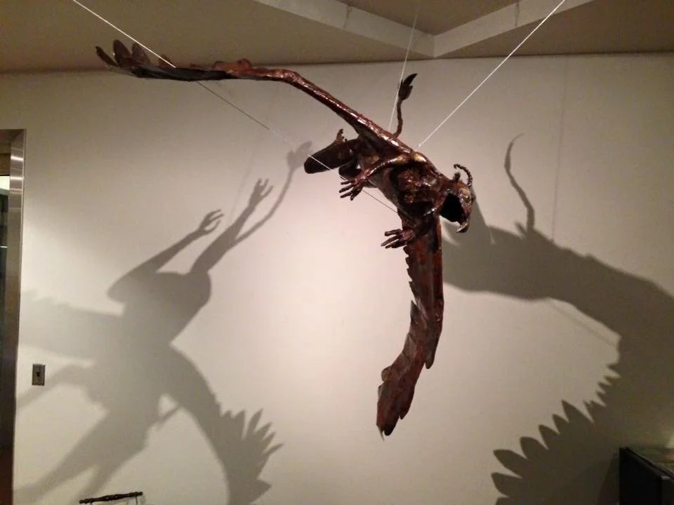 Metal sculpture of a winged creature with a lizard-like body and dragonfly wings, casting large shadows on the wall.