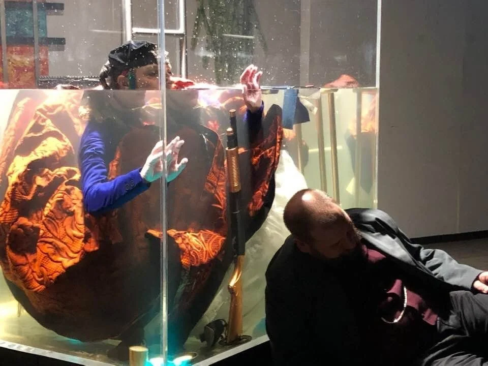 A woman in a blue shirt and headscarf appears to be underwater in a glass tank, with her face and hands pressed against the glass, while a man in a dark suit is lying on the floor beside the tank.