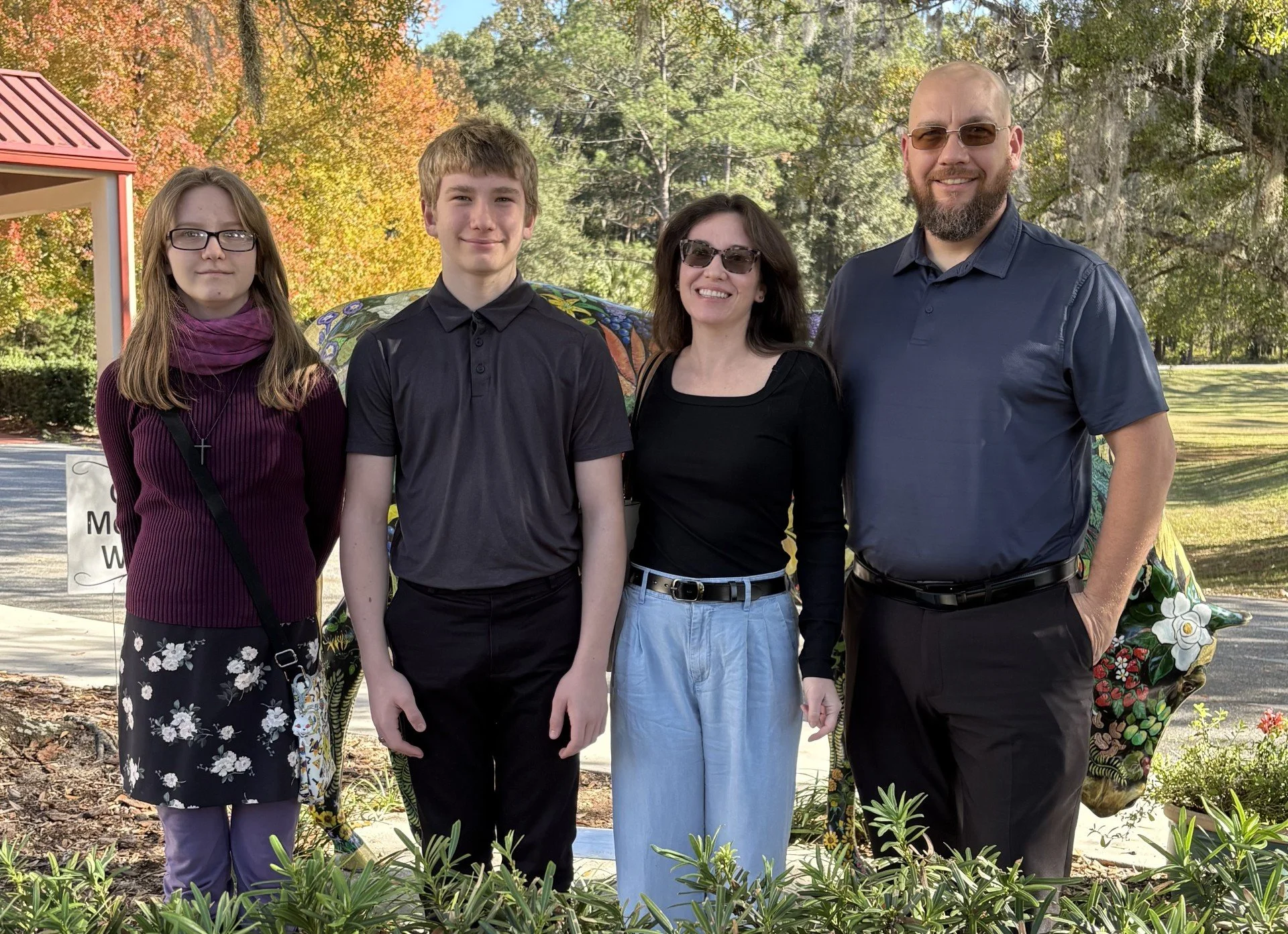 A family of four standing outdoors in front of trees with autumn foliage, smiling at the camera.